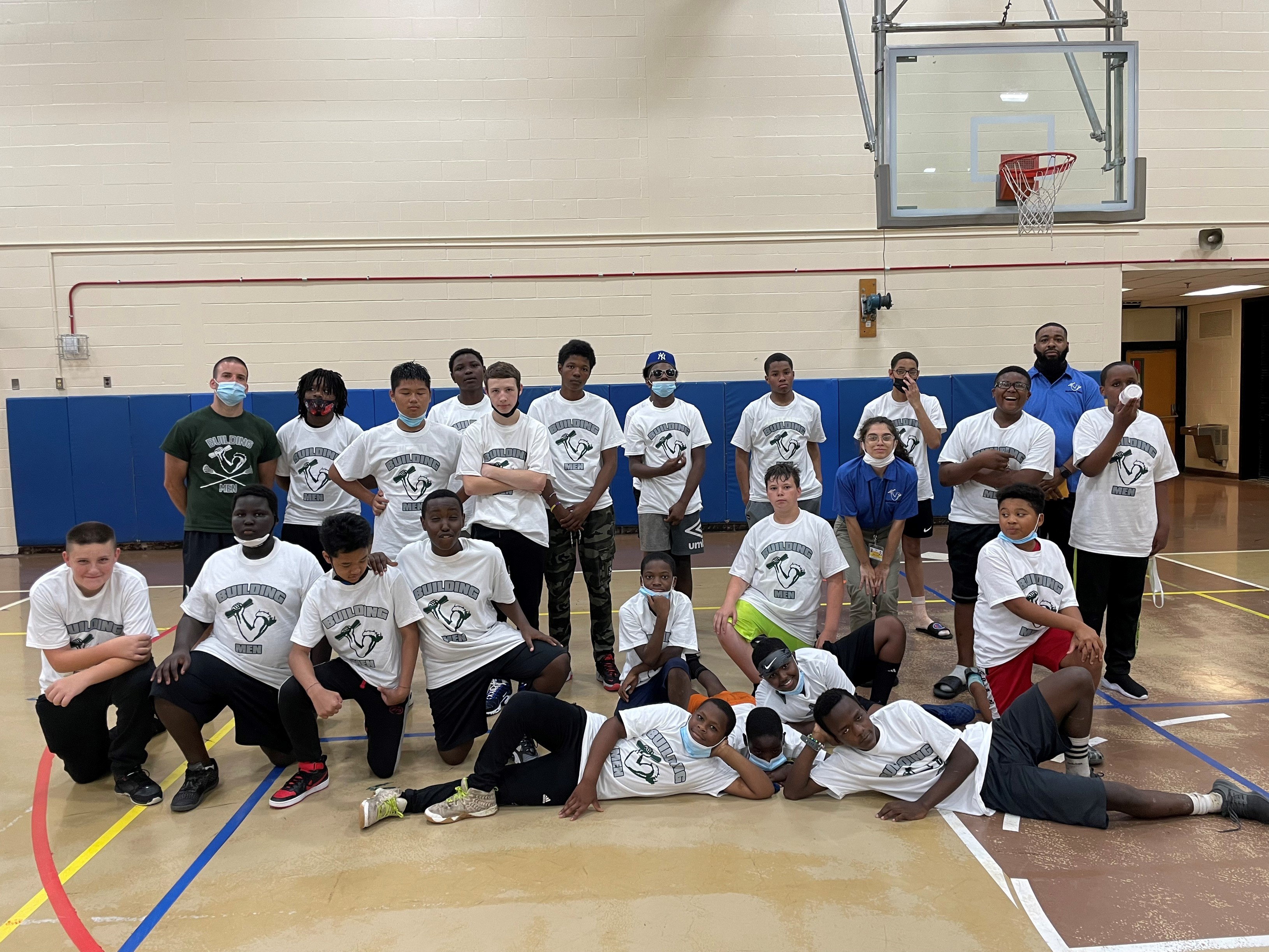 This is a photo of a group of Building Men students posing in a line in their school gym, wearing matching Building Men tshirts.