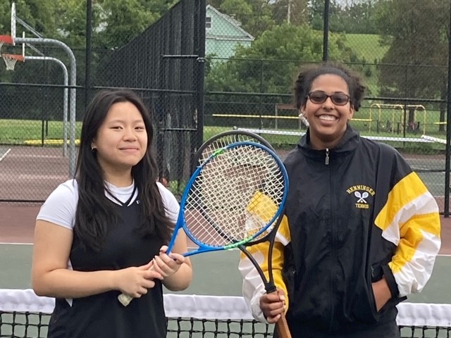 This is a photo of two members of the Henninger girls tennis team, holding their racquets while standing on the tennis court, smiling at the camera.