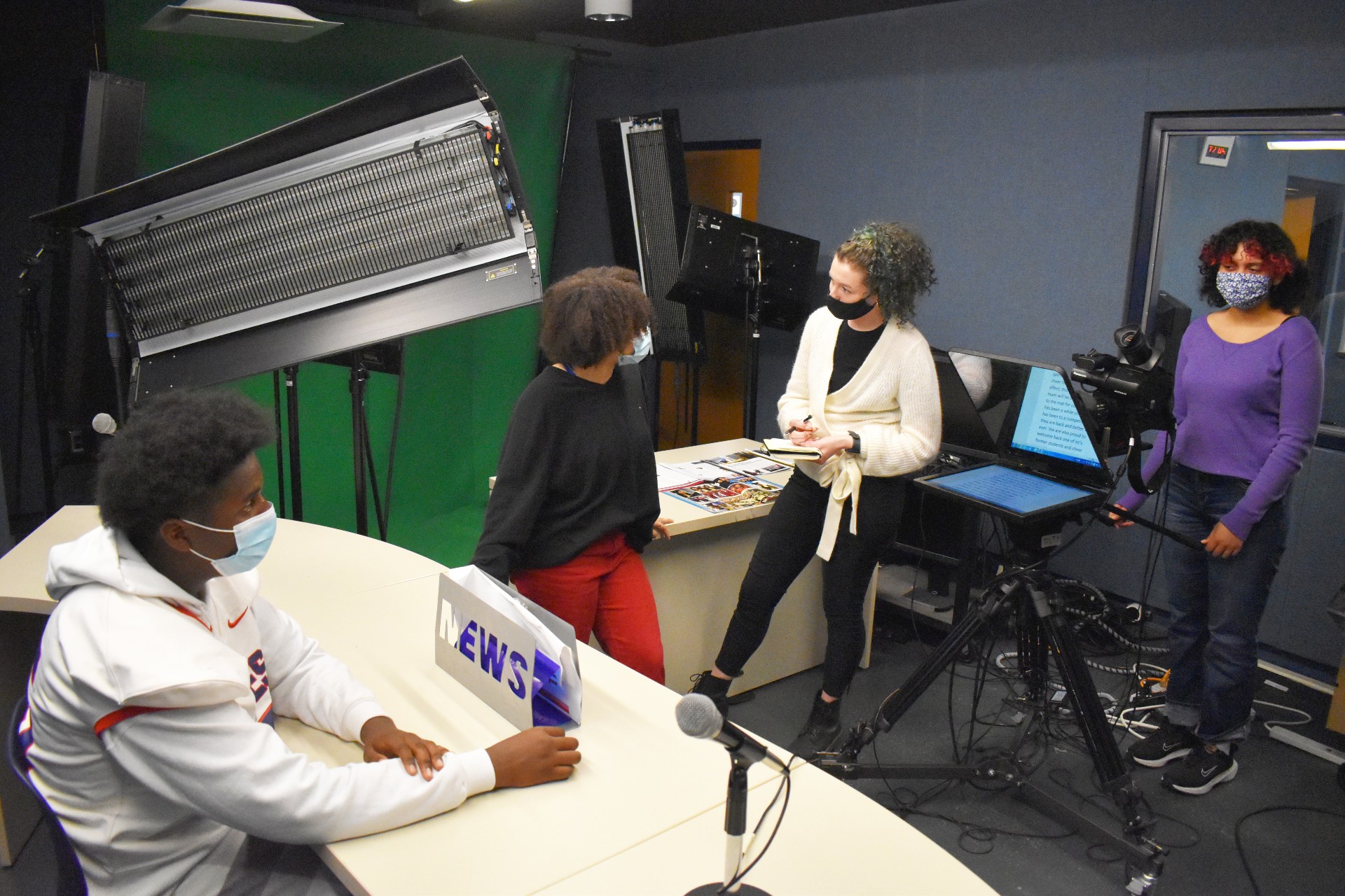 This is a photo of journalism students speaking with their professional mentor, all standing in the ITC media lab, featuring a newsroom setup and news camera.