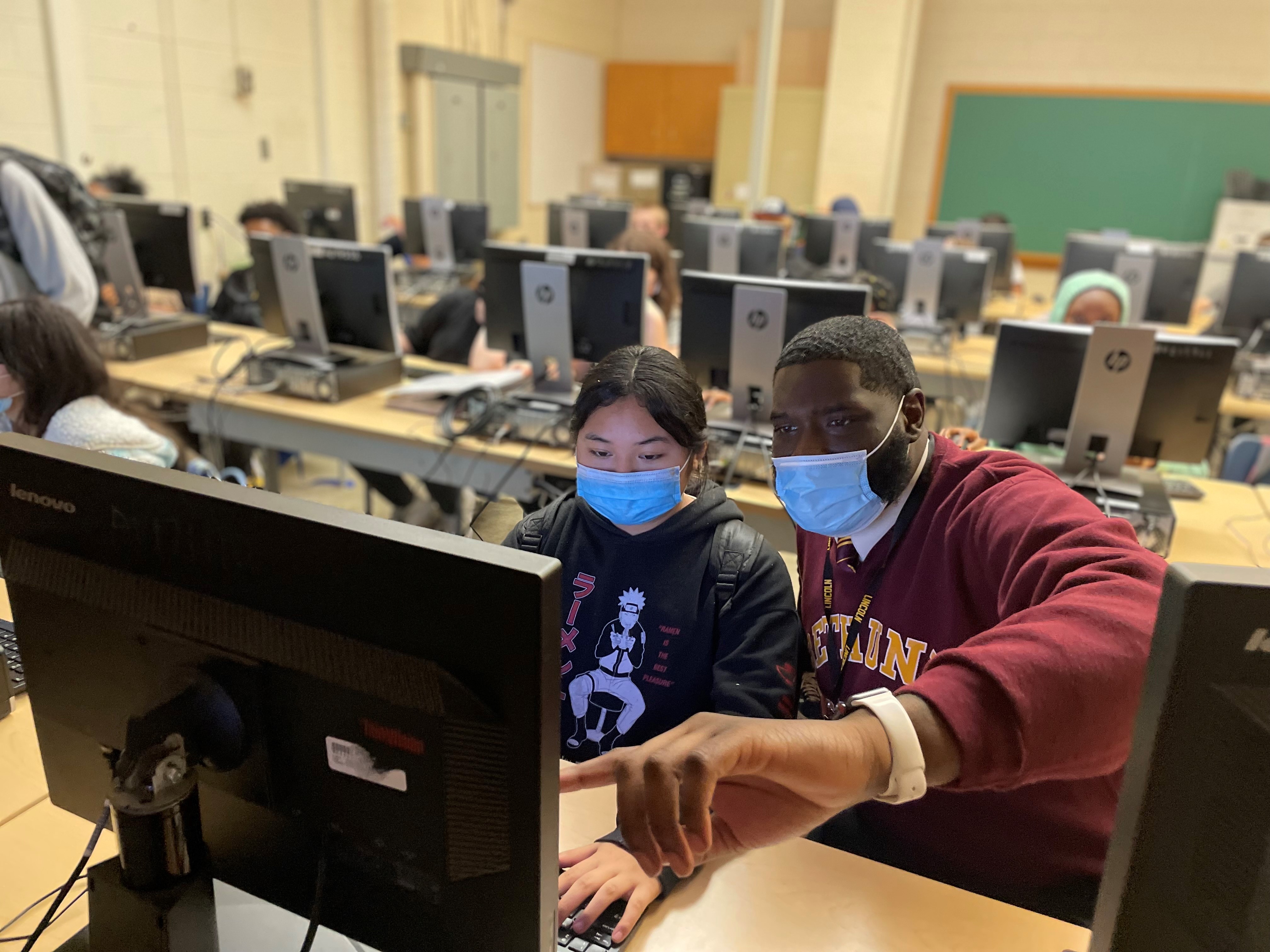 This is a photo of a computer lab, with a female student sitting at her computer, male teacher looking on, pointing to the screen.