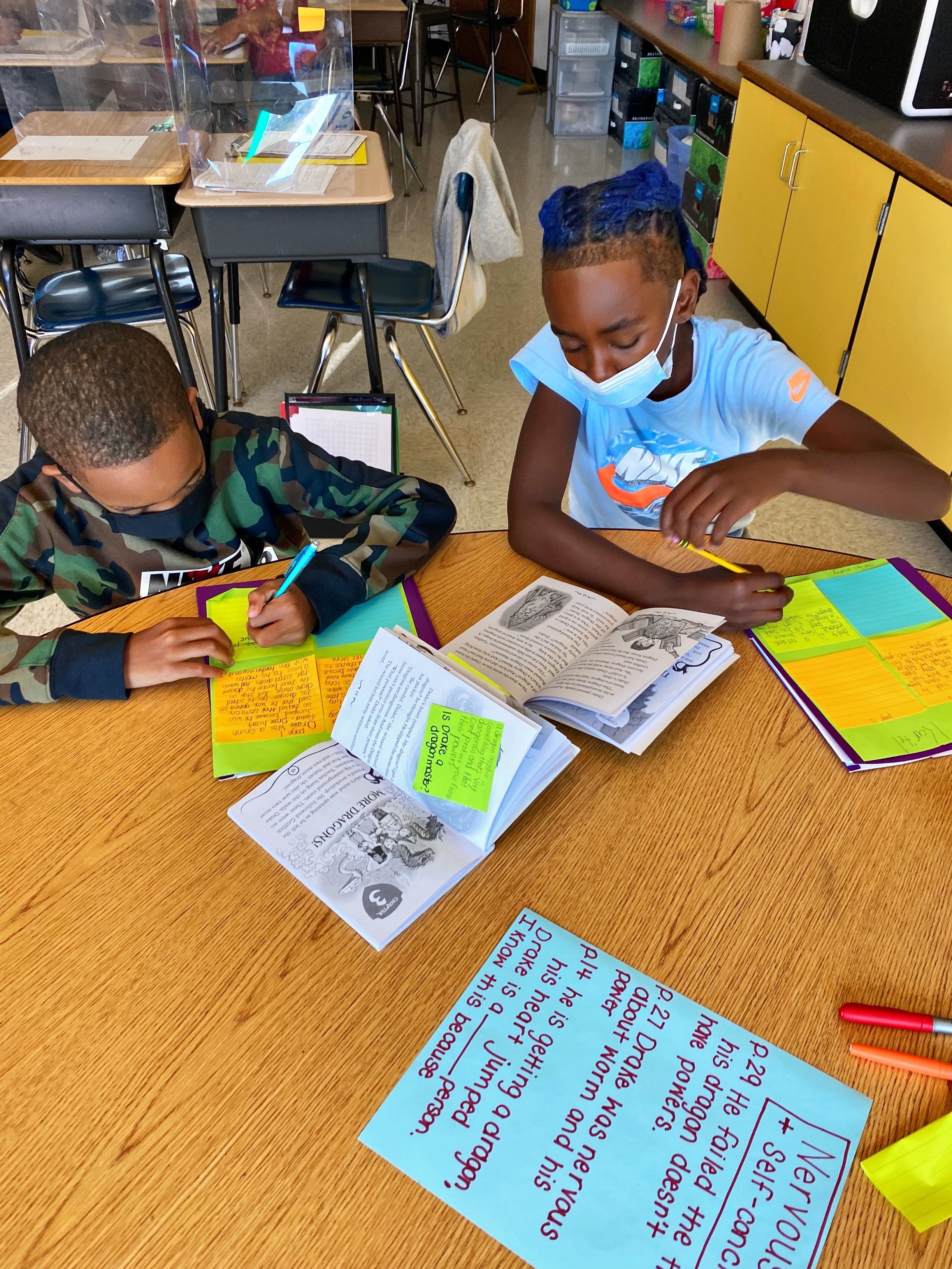 This is a photo of two students sitting at desks looking over books covered with post-it notes.