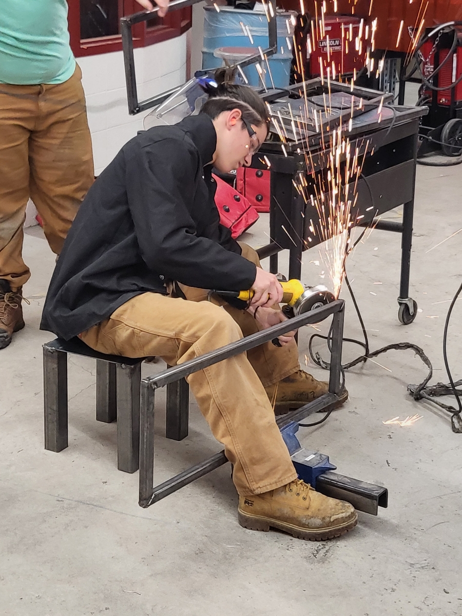 This is a photo of a male student wearing PPE using a welding torch to weld a metal basketball cart together.