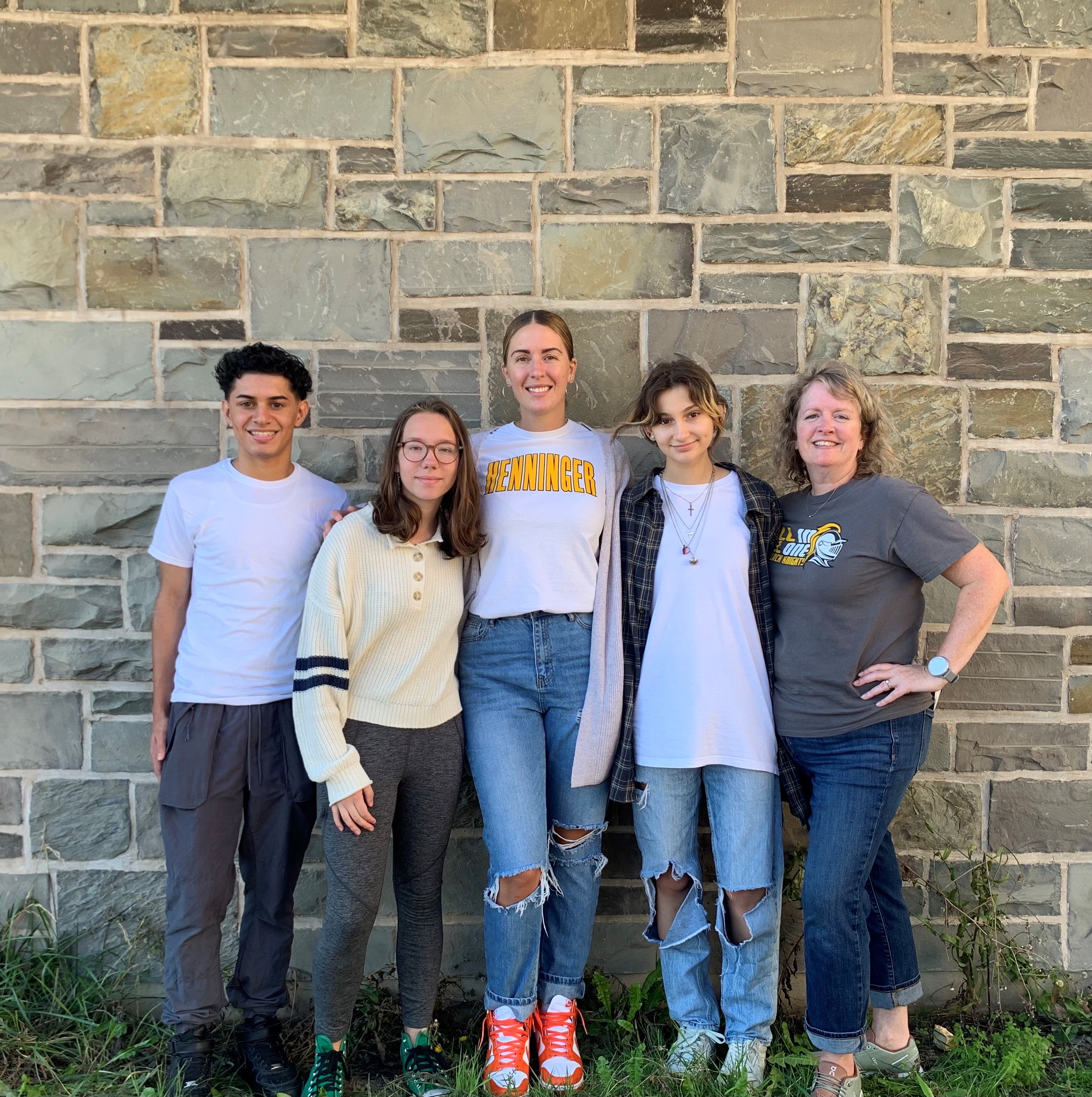 This is a photo of three Henninger students, an alumna, and an art teacher, standing in a line in front of a stone wall smiling at the camera.