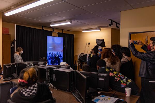This is a photo showing a classroom of students crowded around a large video monitor, where students located in South Africa are displayed smiling back.