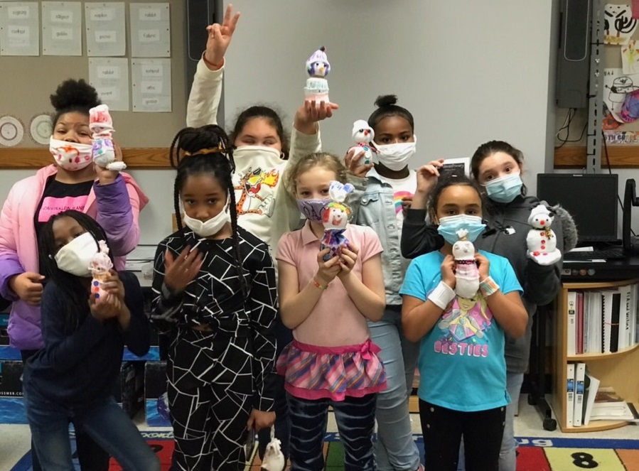This is a photo of a group of girls standing together, wearing masks, each holding up a snowman they made out of a sock.
