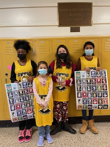 This is a photo of four Huntington students, each wearing a yellow jersey that says 'Arria' standing in front of lockers holding two posters showing photos of the women who will be honored during the Women's Empowerment Draft event.