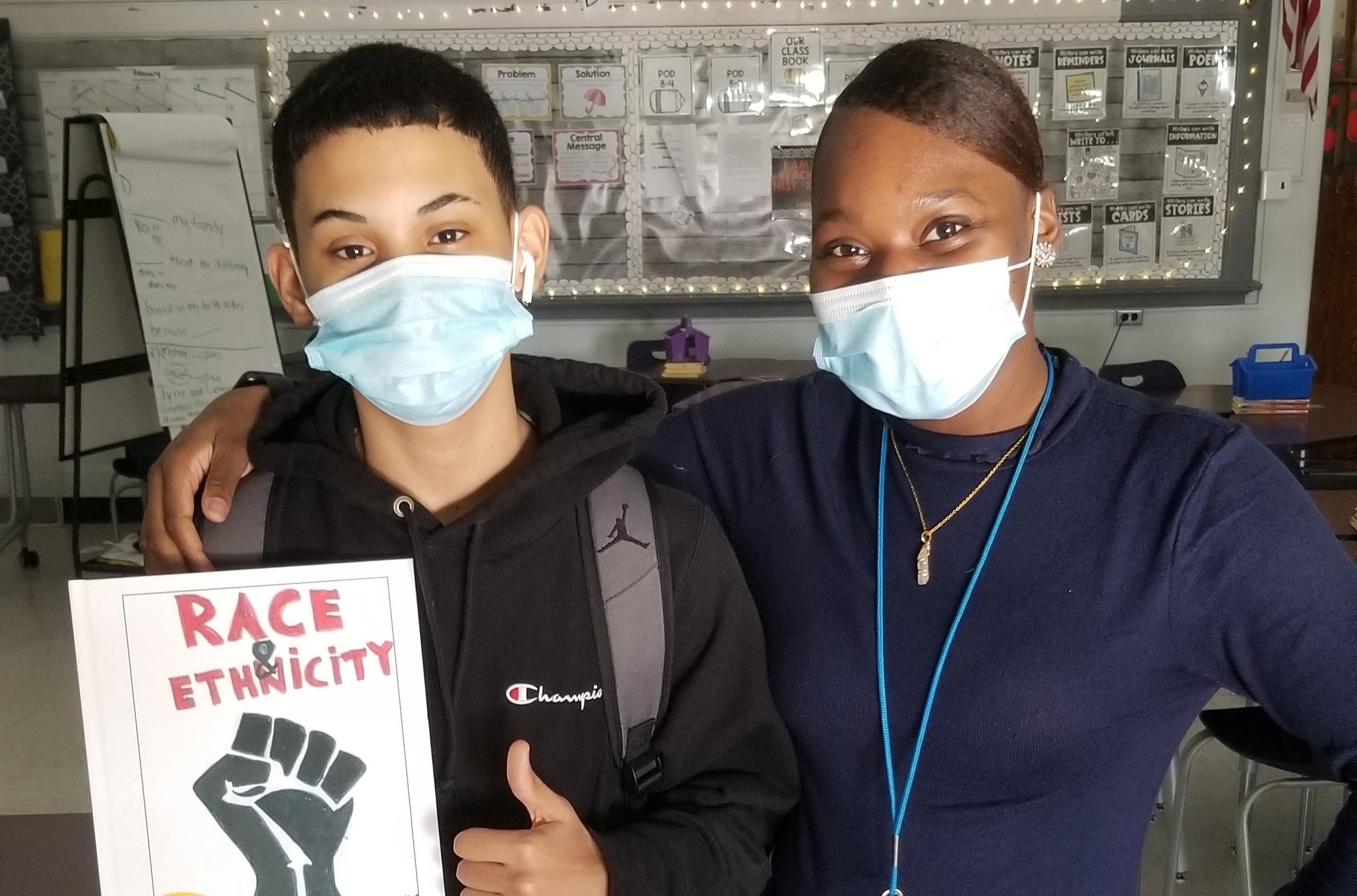 This is a photo of a male student holding a book titled 'Race & Ethnicity' standing next to his teacher.