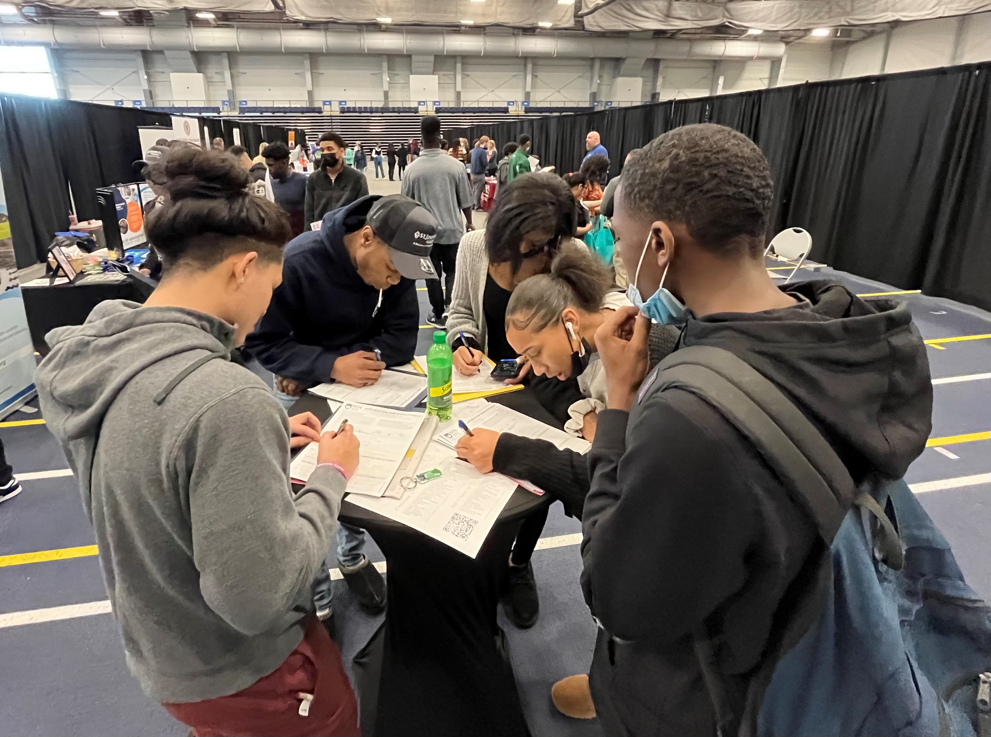 This is a photo of several students crowding around a round table, each completing a job application at a CTE Career Fair.