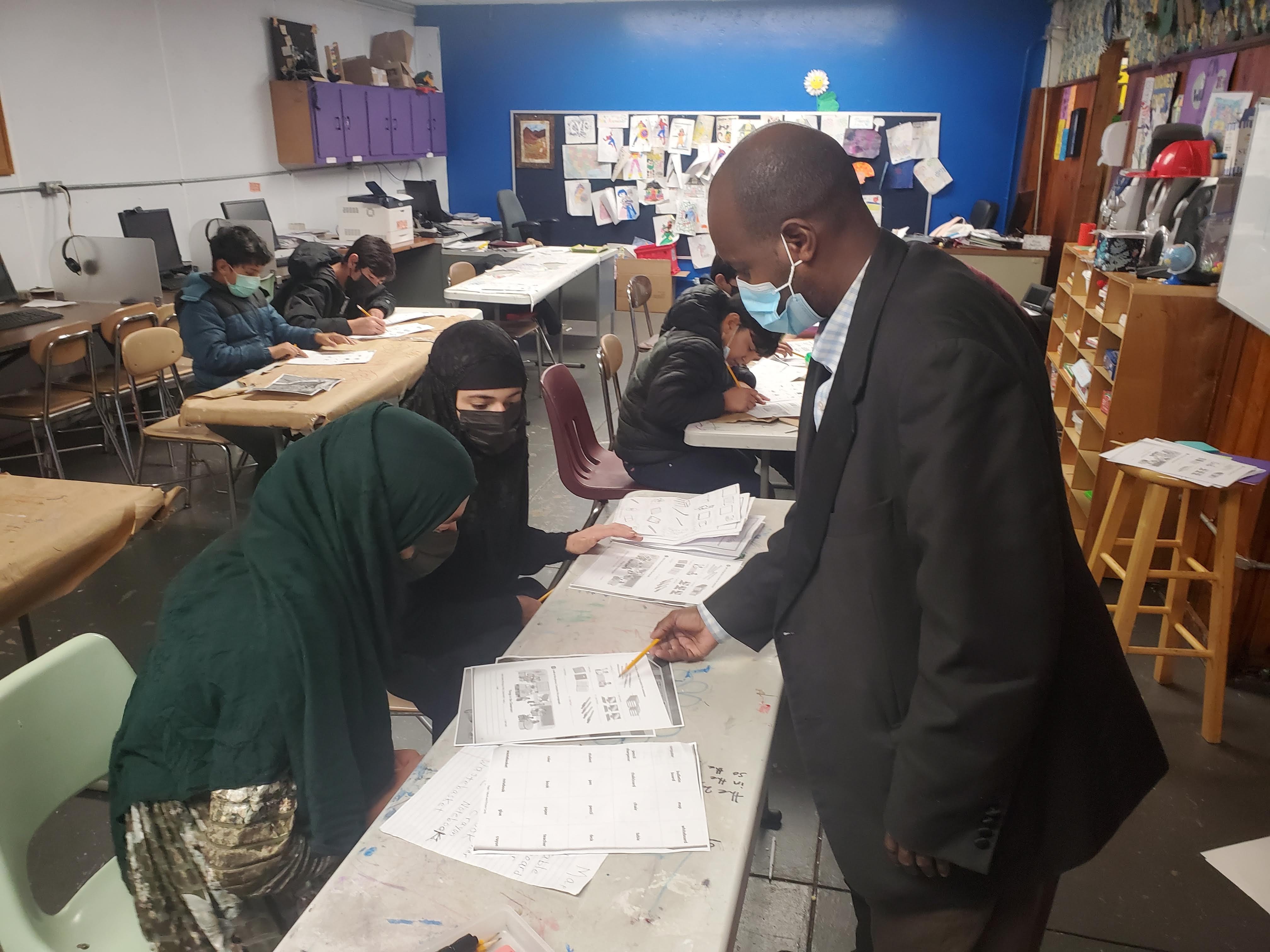 This is a photo of two students sitting at a desk looking at a paper, while the teacher stands in front of them, pointing to the paper.