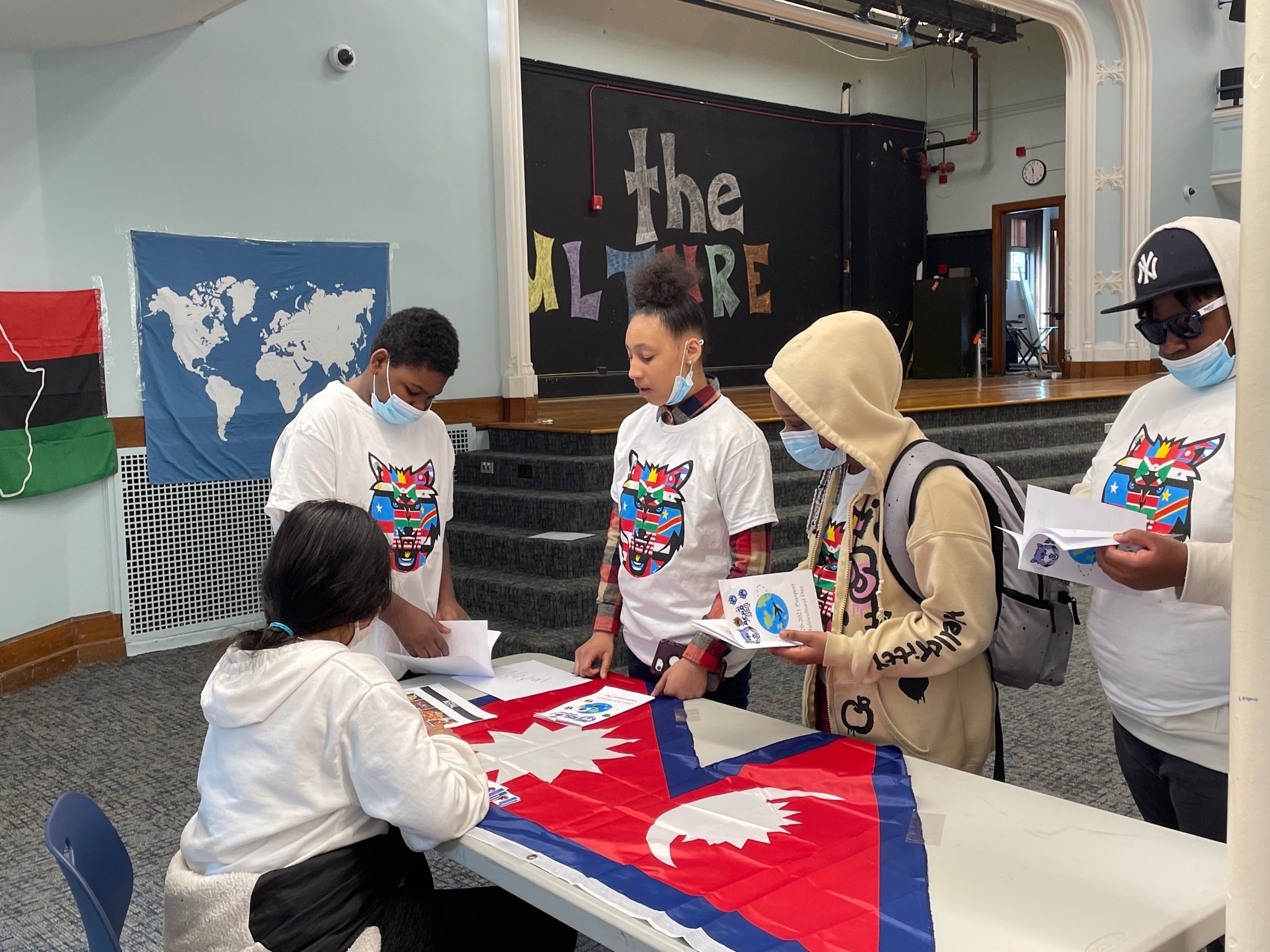 This is a photo of four students standing at a table, one sitting, all looking at the red flag laid on the table.