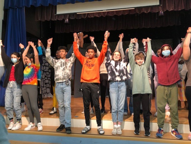 This is a photo of the Ed Smith students performing in the spring musical, standing on the steps of the school stage holding hands above their heads as they take a group bow during a dress rehearsal.