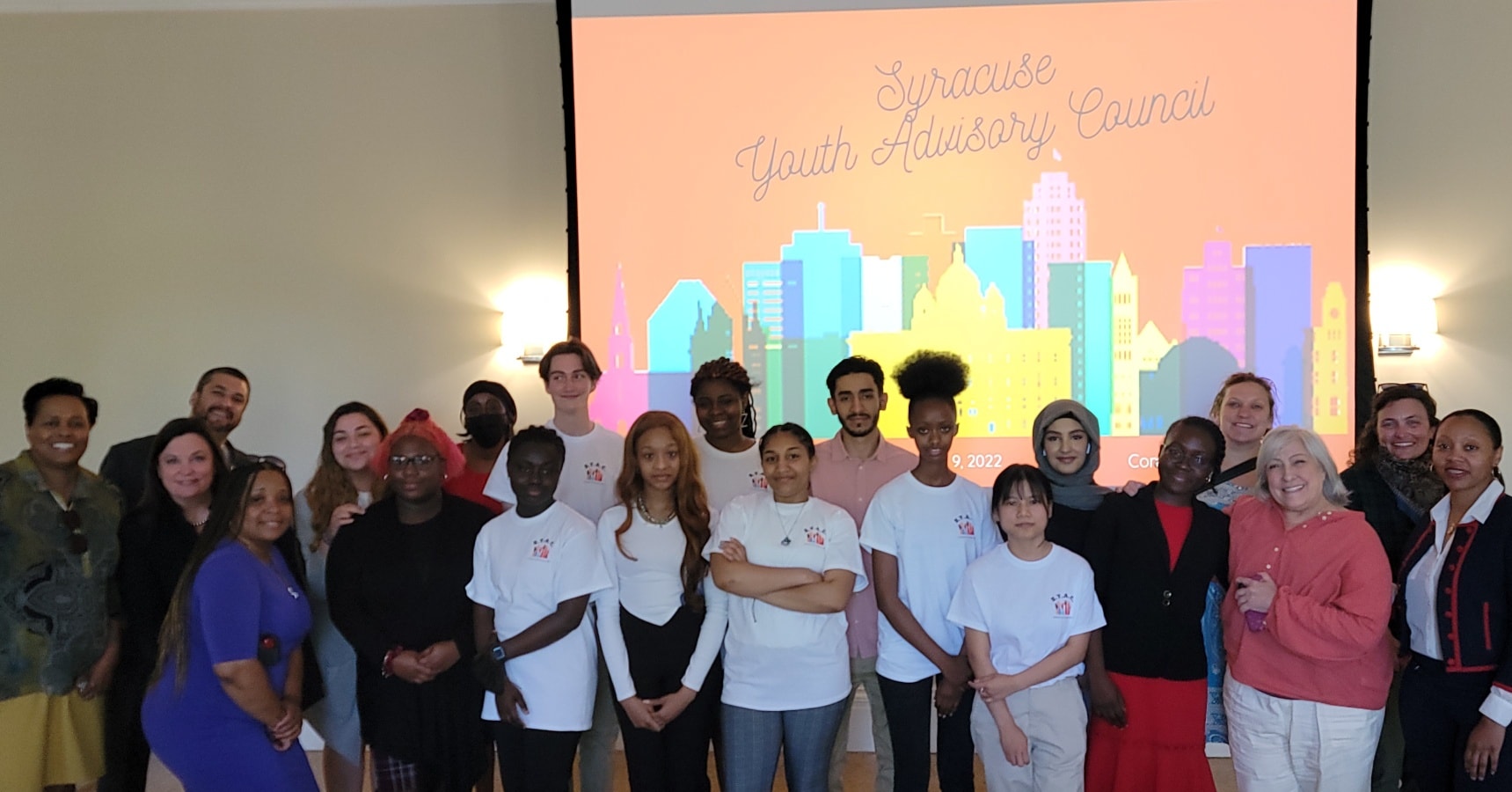 This is a group photo of all members of the Syracuse Youth Advisory Council, standing in front of a slide projection that reads 'Syracuse Youth Advisory Council.'