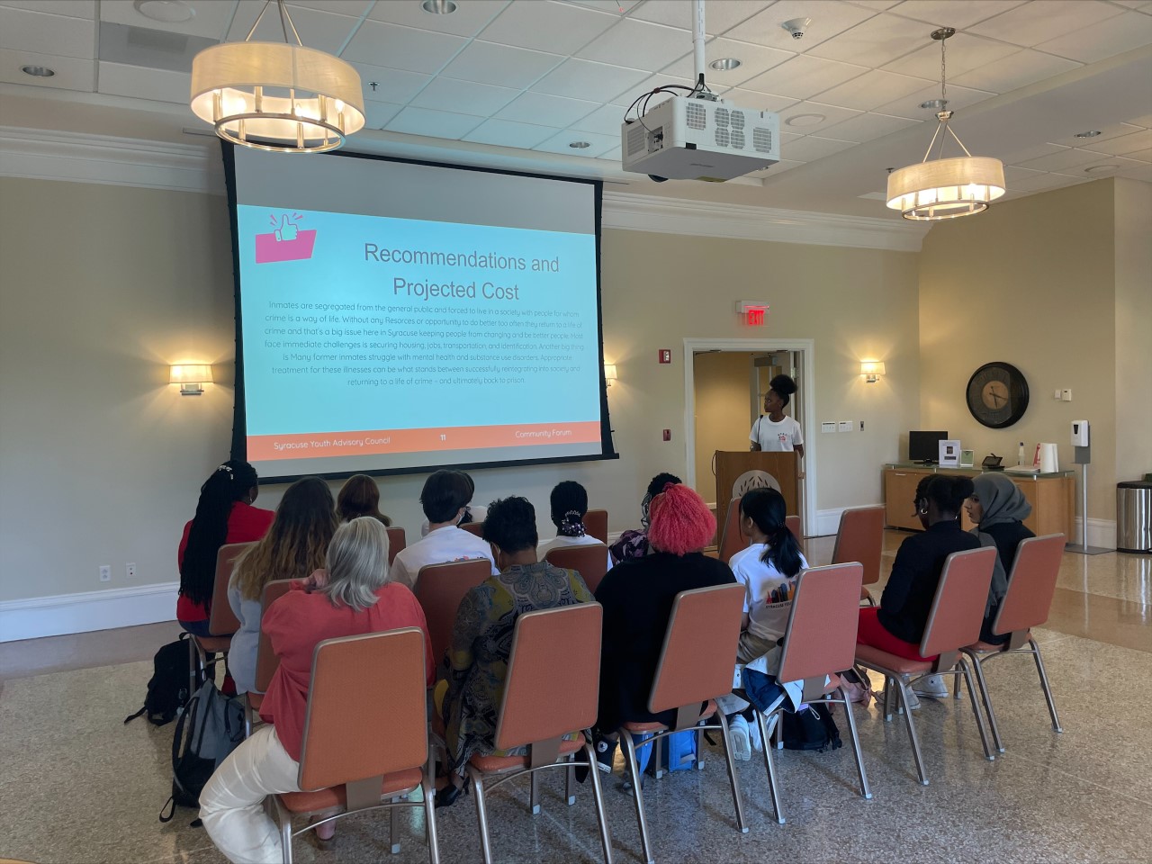 This is a photo of a PSLA at Fowler student standing at a podium next to a slide projection presenting to an audience seated in rows of chairs.