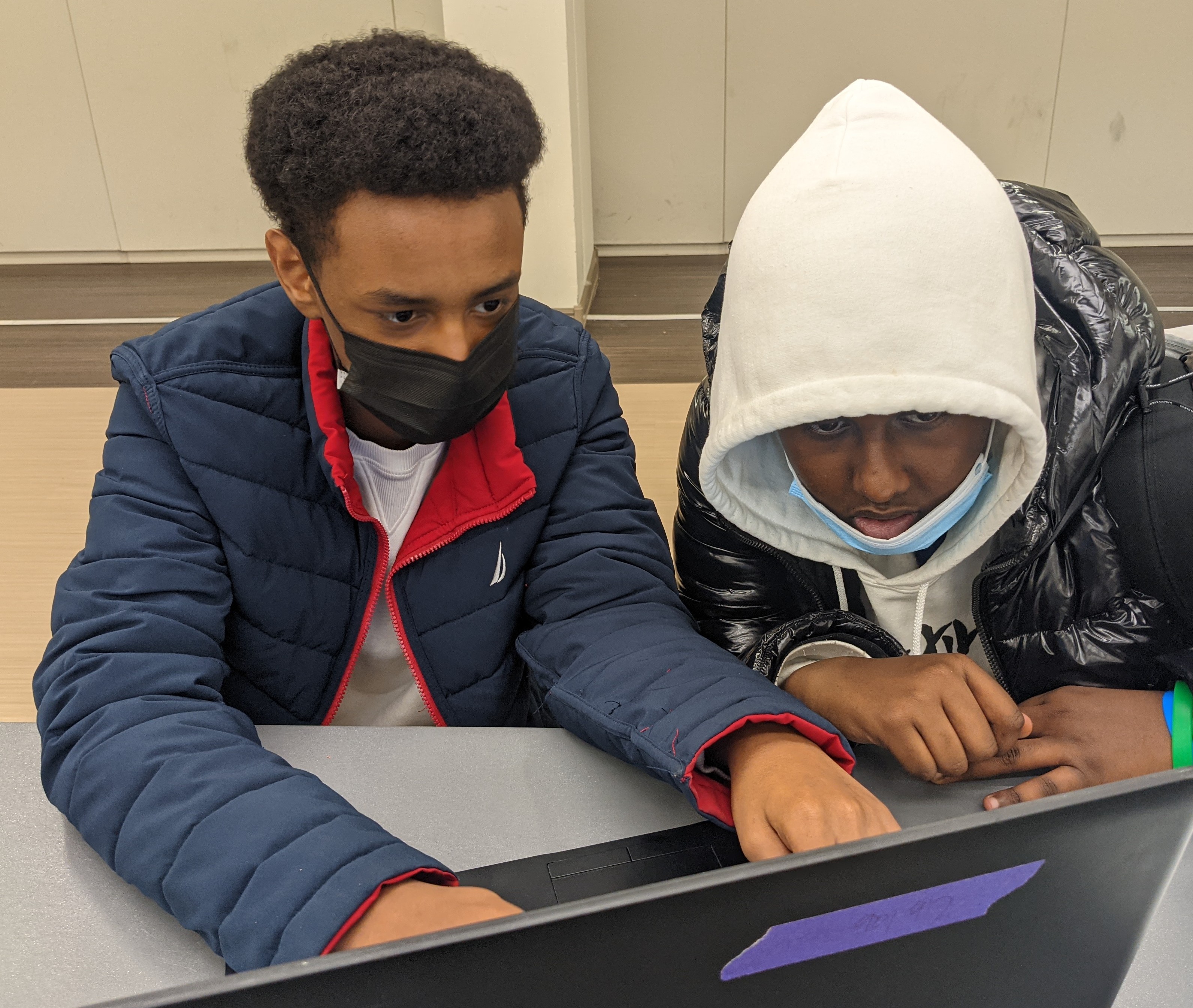 Two male students wearing masks sit at a computer, typing. 