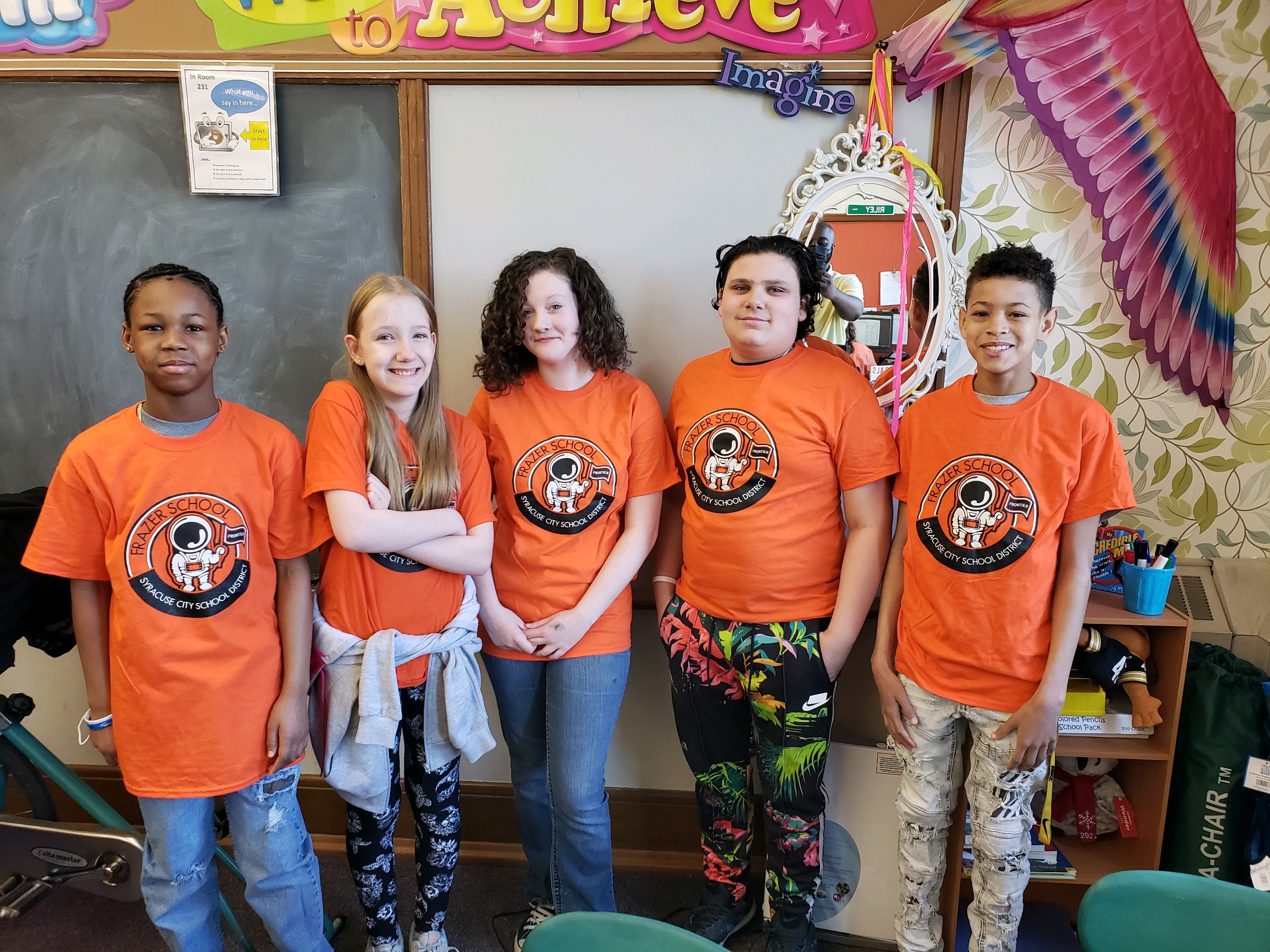 This is a photo of five Frazer students standing in a line in front of a chalkboard, all wearing matching orange Frazer tshirts.