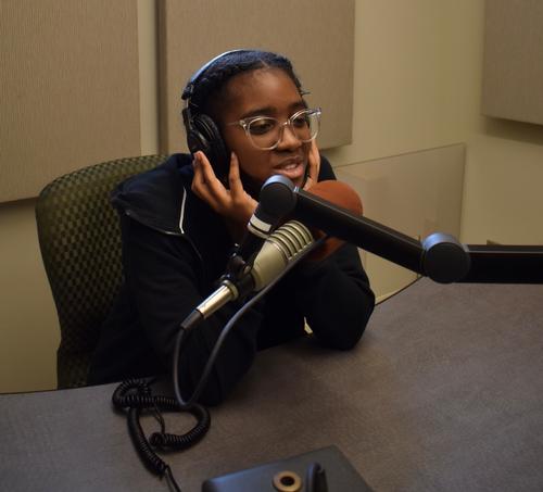 This is a photo of a girl sitting at a microphone, wearing headphones, in a recording studio at Syracuse University.