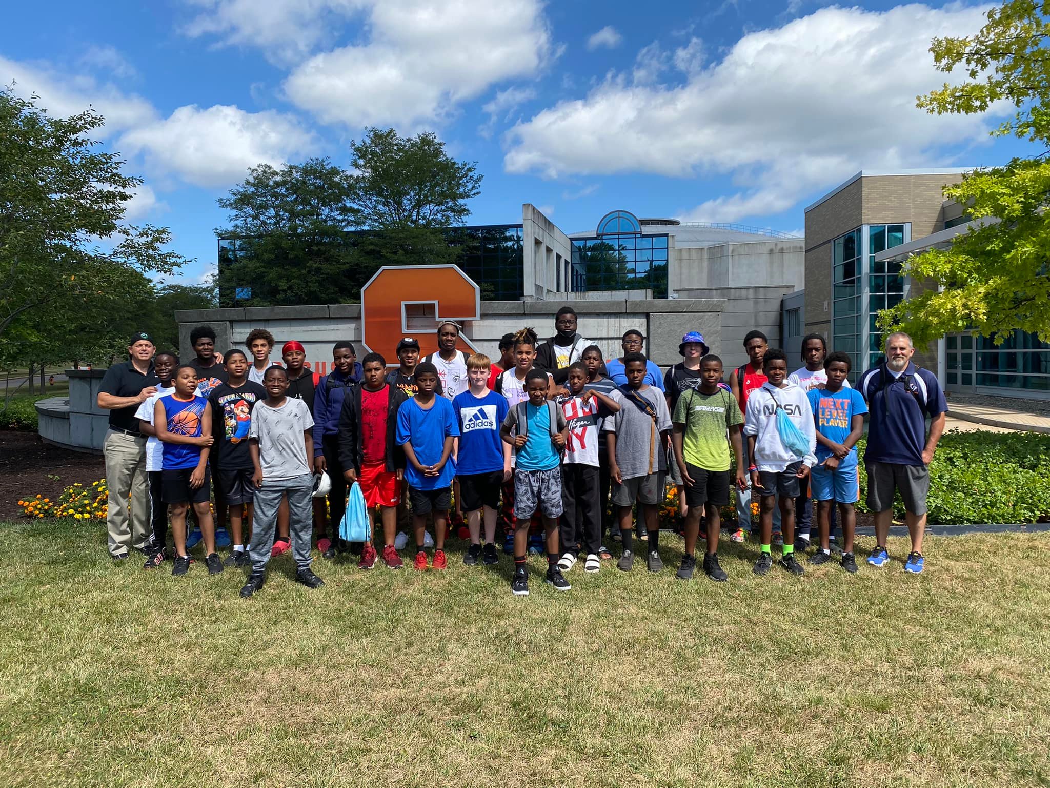 This is a photo of a large group of Building Men students standing in front of an orange S sculpture on the Syracuse University campus.