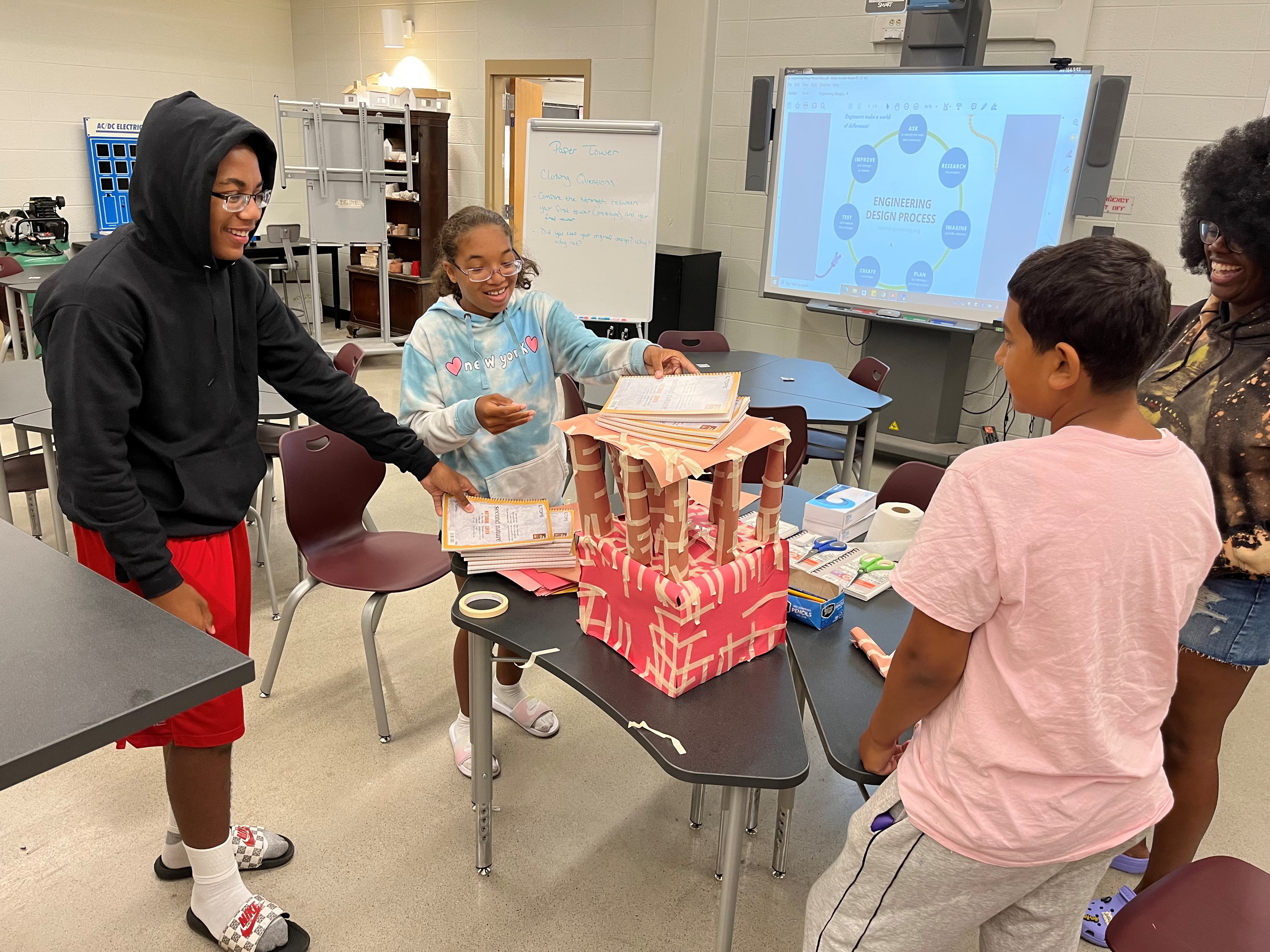 This is a photo of four students smiling as they stand around a tabletop tower made out of paper.