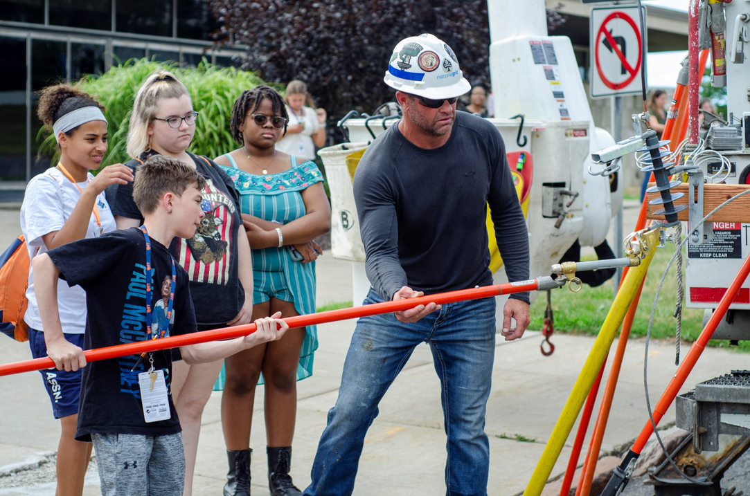 This is a photo of four students looking on as a man in a hard hat demonstrates something with an electrical truck.