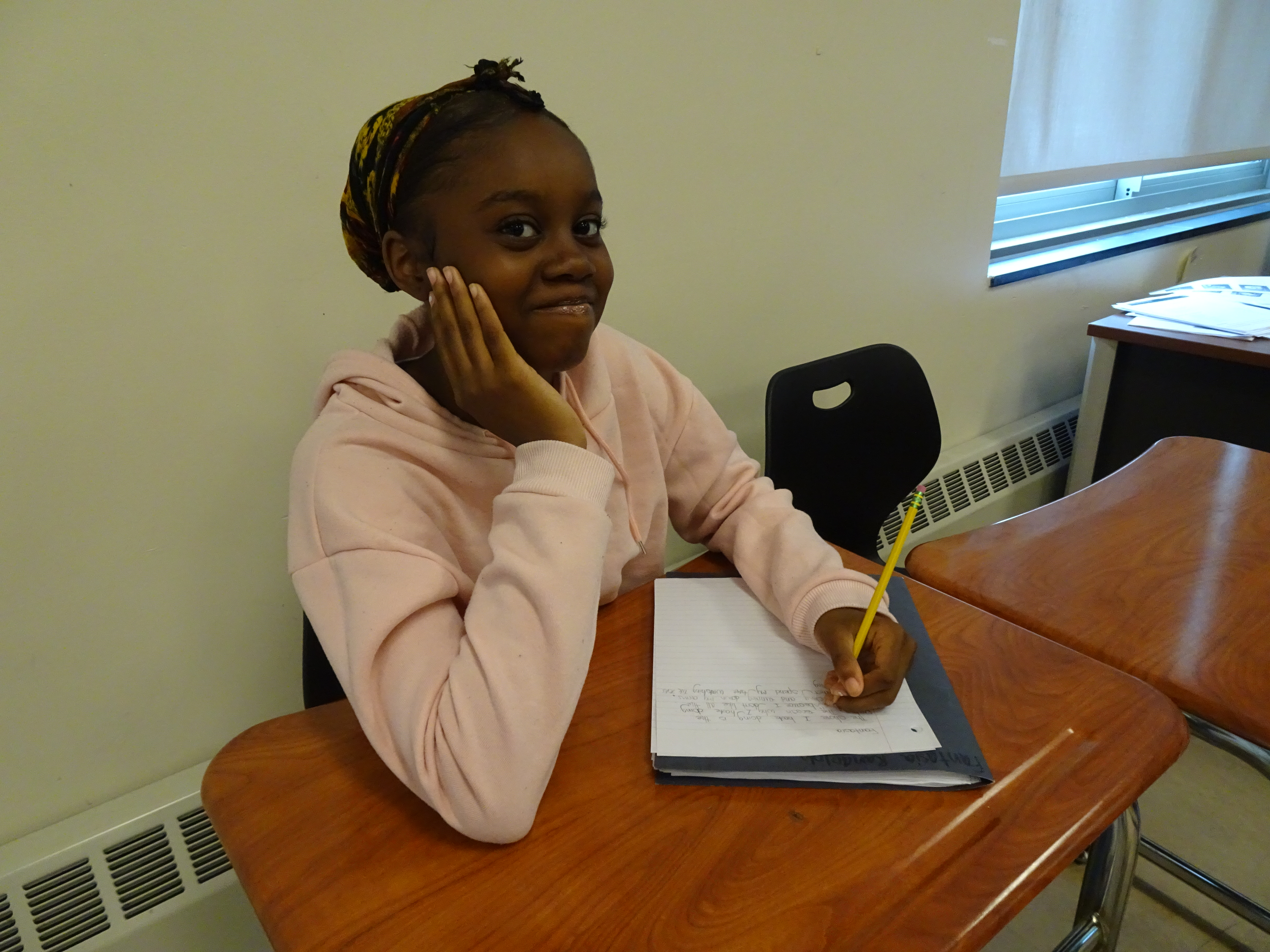 This is a photo of a girl in a pink sweater sitting at a desk, writing in a notebook and smiling at the camera.