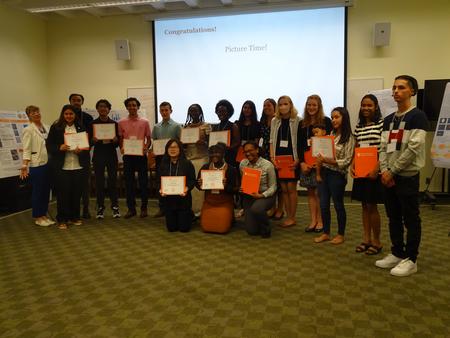 This is a photo of SCSD students who interned at Syracuse University, standing in two rows in a presentation room at SU holding certificates of completion.