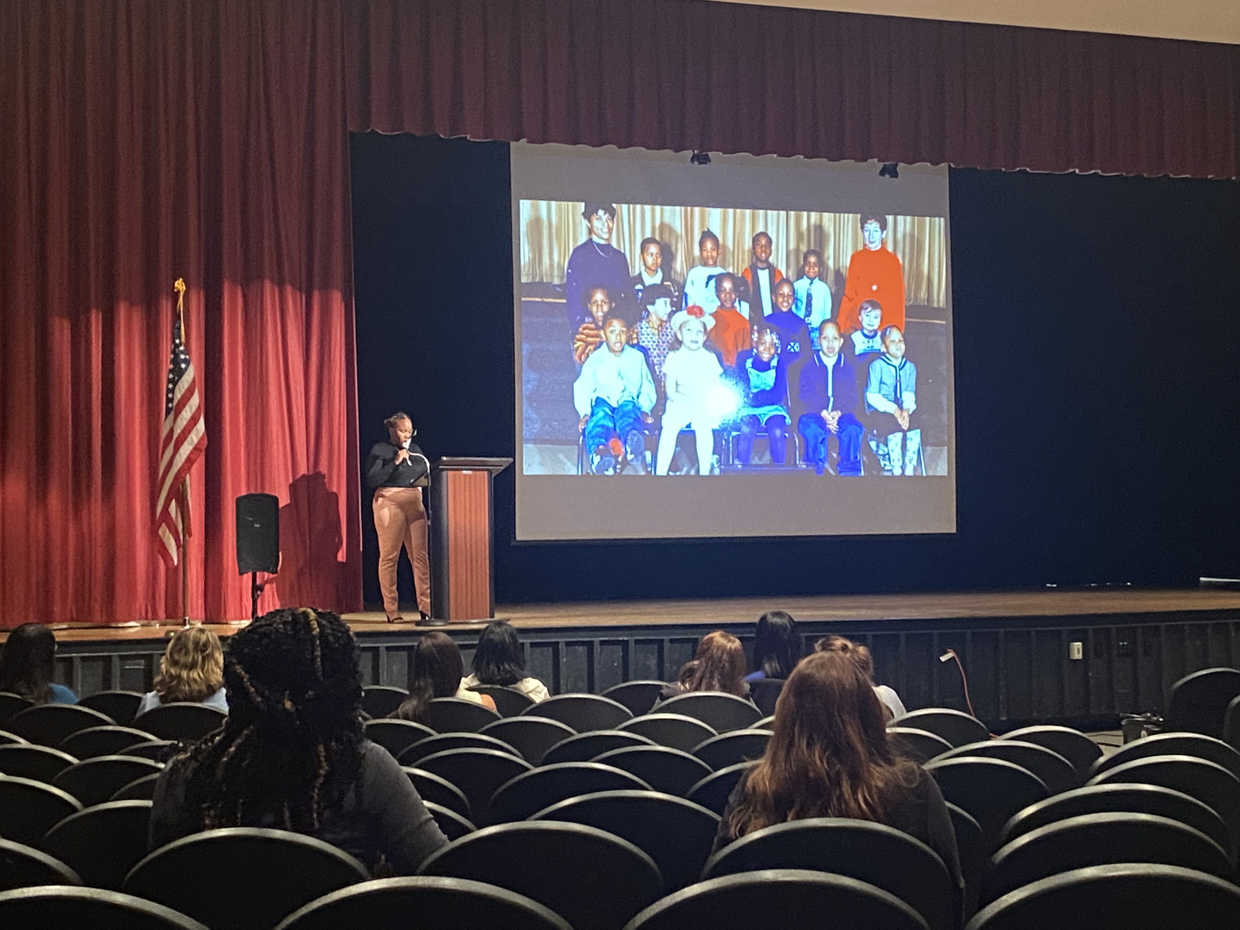 This is a photo of a woman standing on a stage at a podium, with a photo displayed on a large screen to her right.