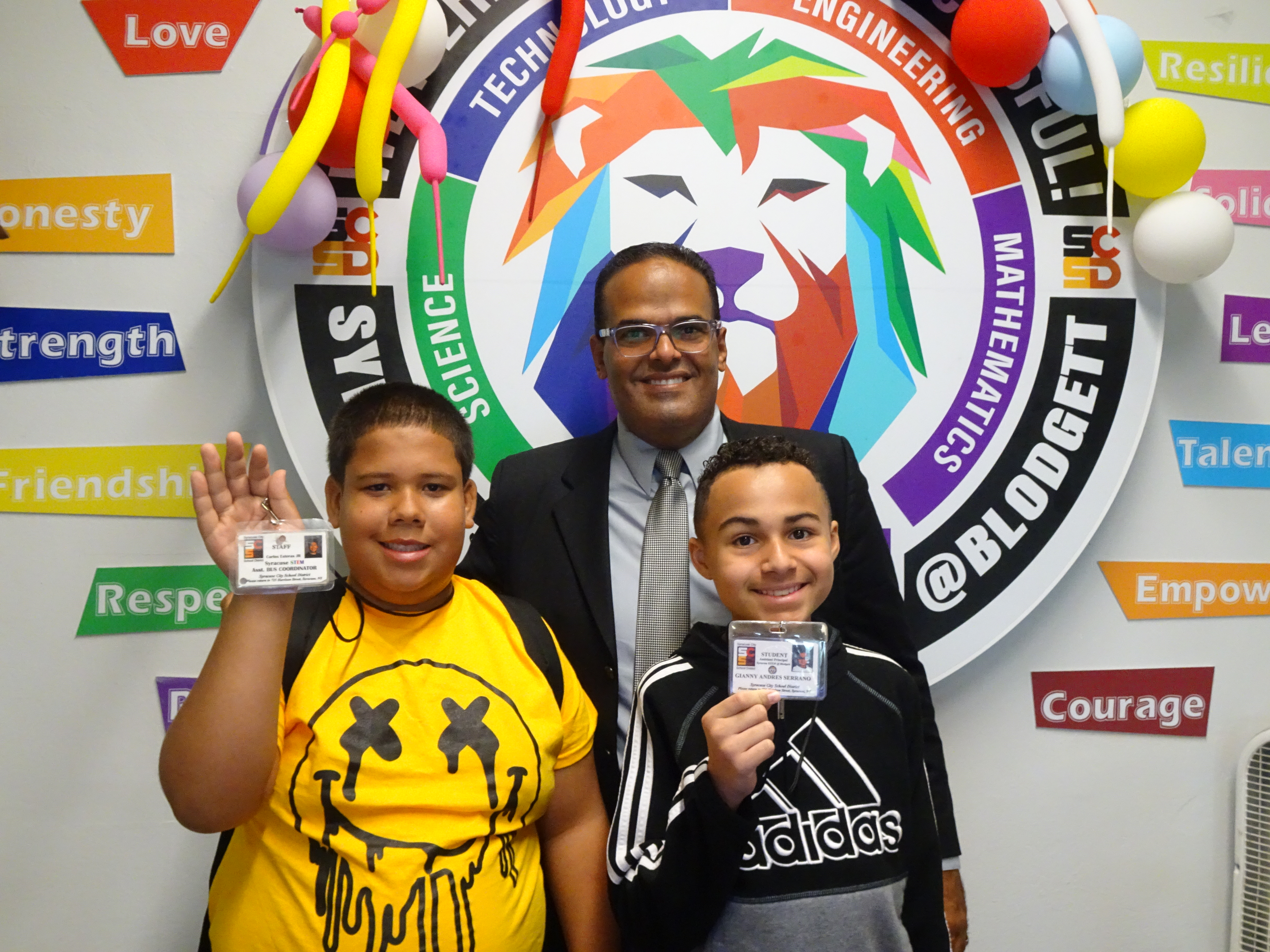 This is a photo of Syracuse STEM at Blodgett Principal Harry Valentin standing with two student administrators, each holding their own ID badge, in front of the school logo hanging on a wall.