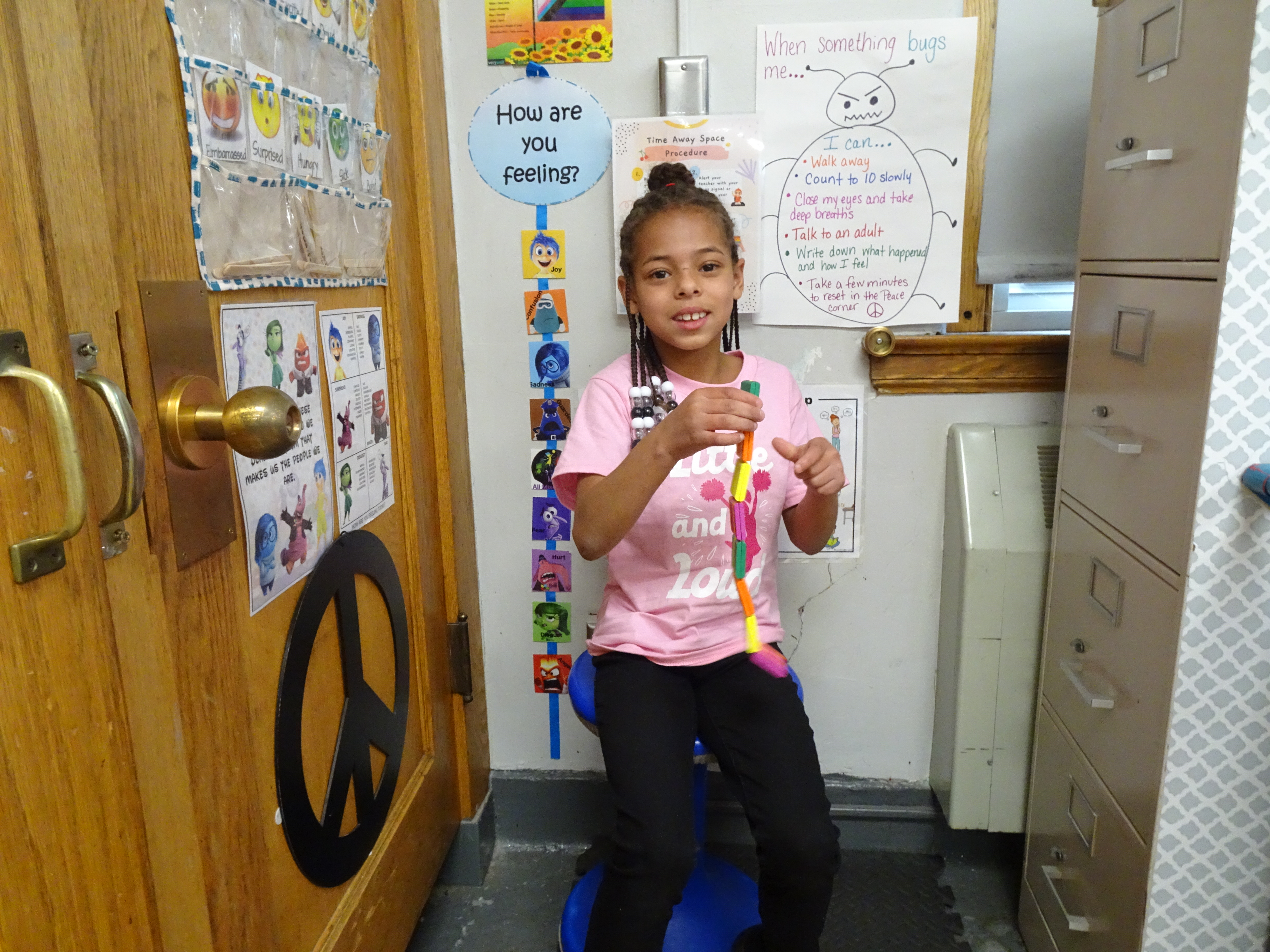 This is a photo of a girl sitting on a chair in a corner of her classroom, playing with a fidget toy.