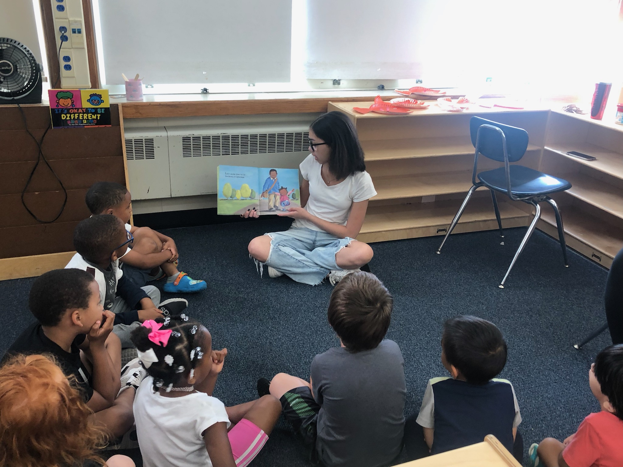 This is a photo of a high school student reading to a group of younger students, sitting in a circle around her as she holds up a book she is reading to them.