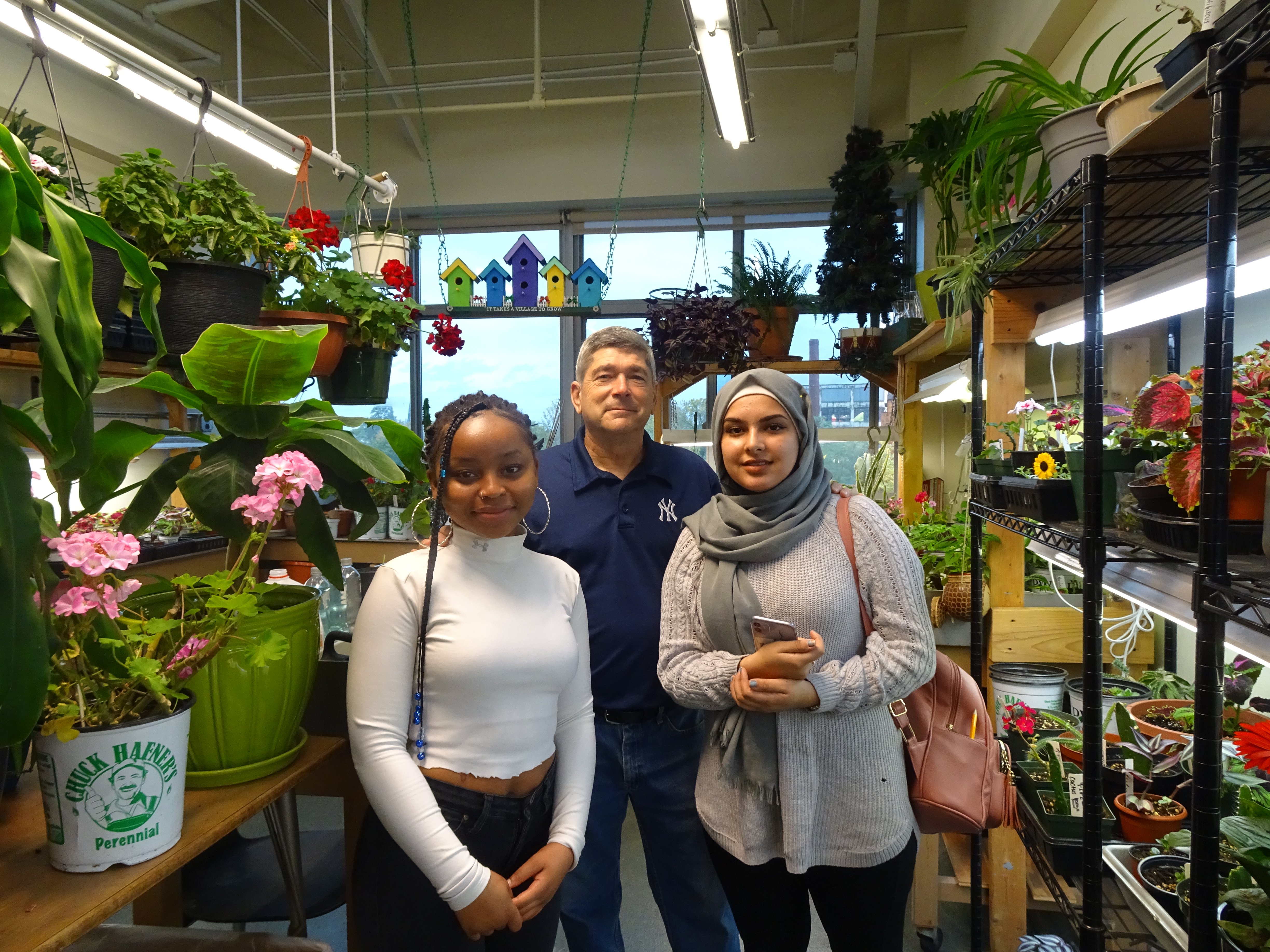 This is a photo of two students and volunteer Dave Sawyer standing in the aisle of their school's greenhouse, surrounded by plants.