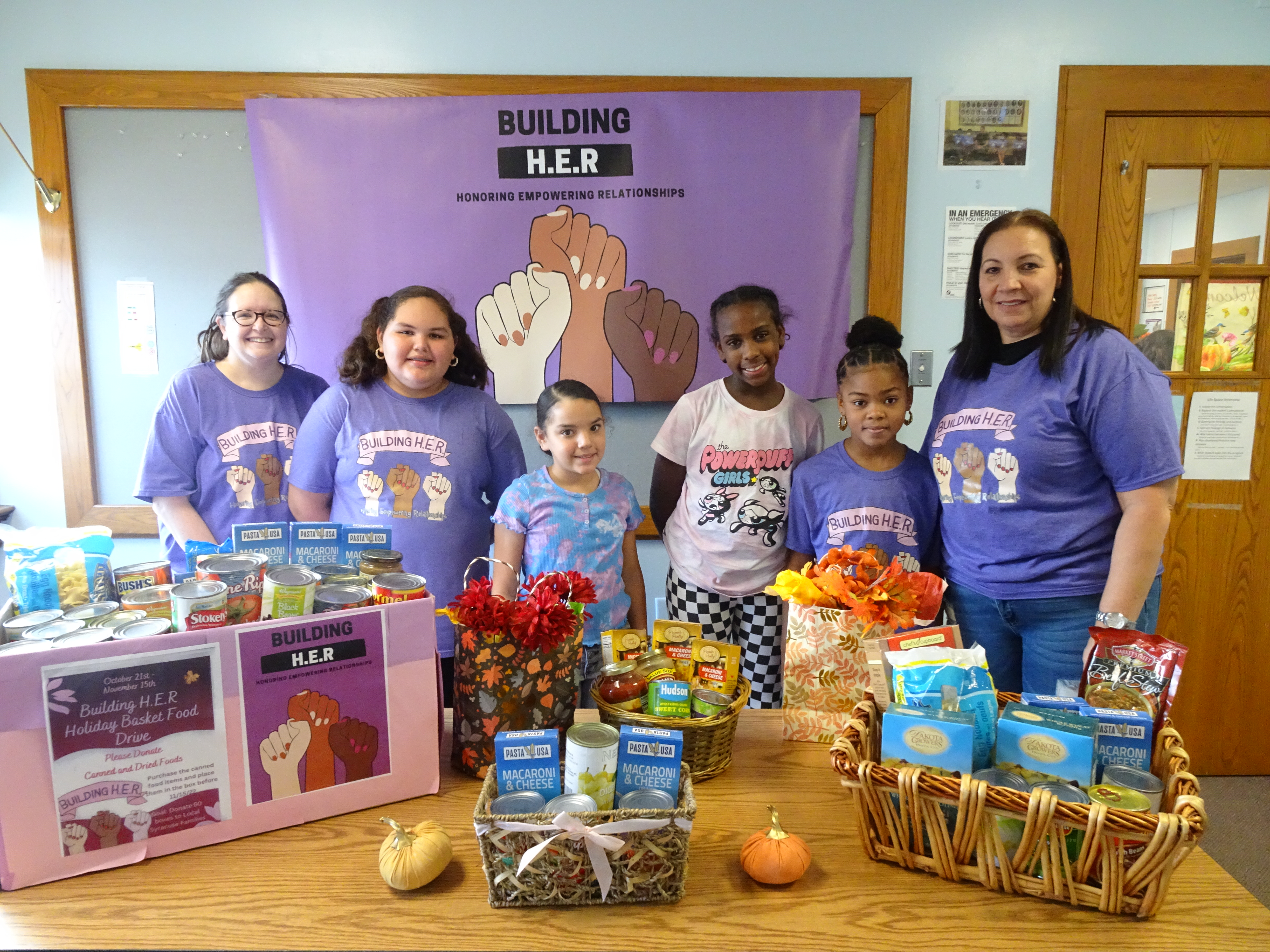 This is a photo of a group of Building HER students standing behind a table with Thanksgiving food baskets on it.