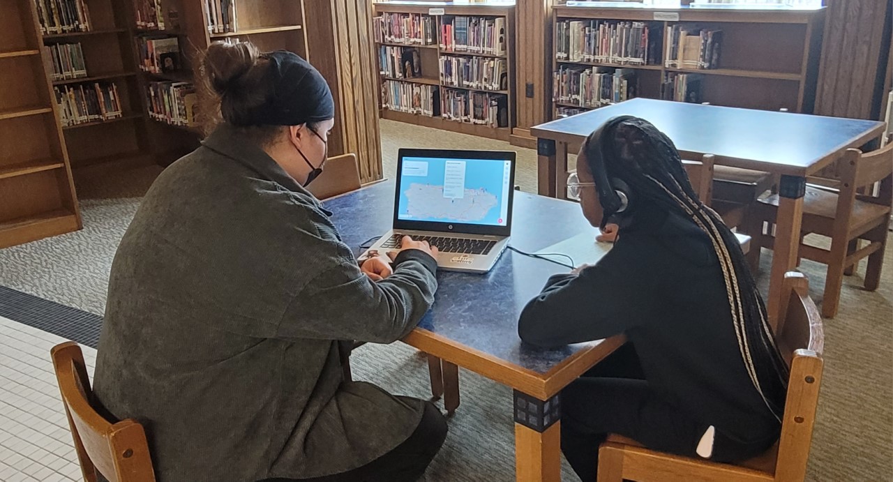 This is a photo of a teacher and a student sitting at a desk together, looking over a computer screen.