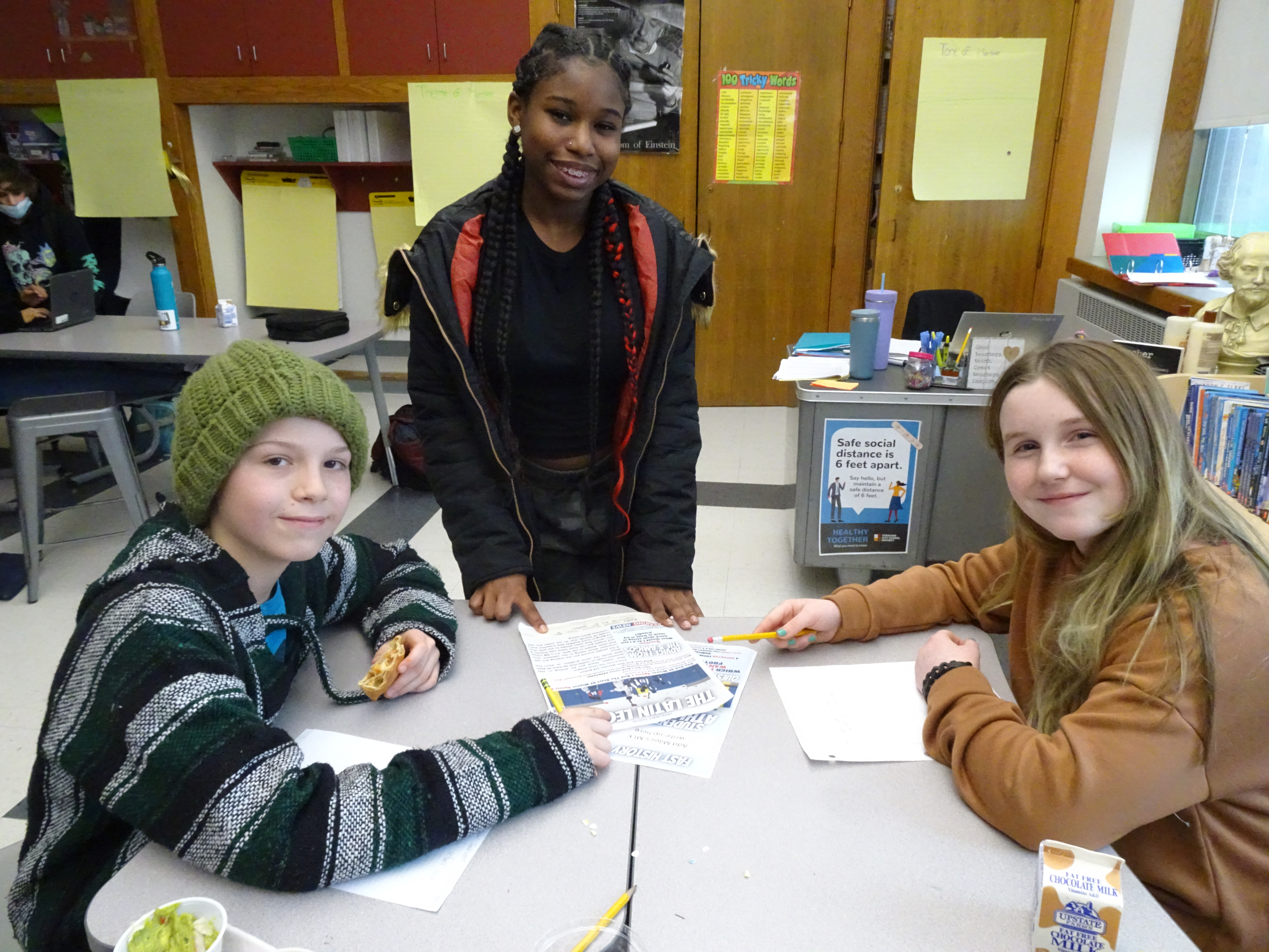This is a photo of three students standing around a desk, pointing to their newsletter while smiling at the camera.