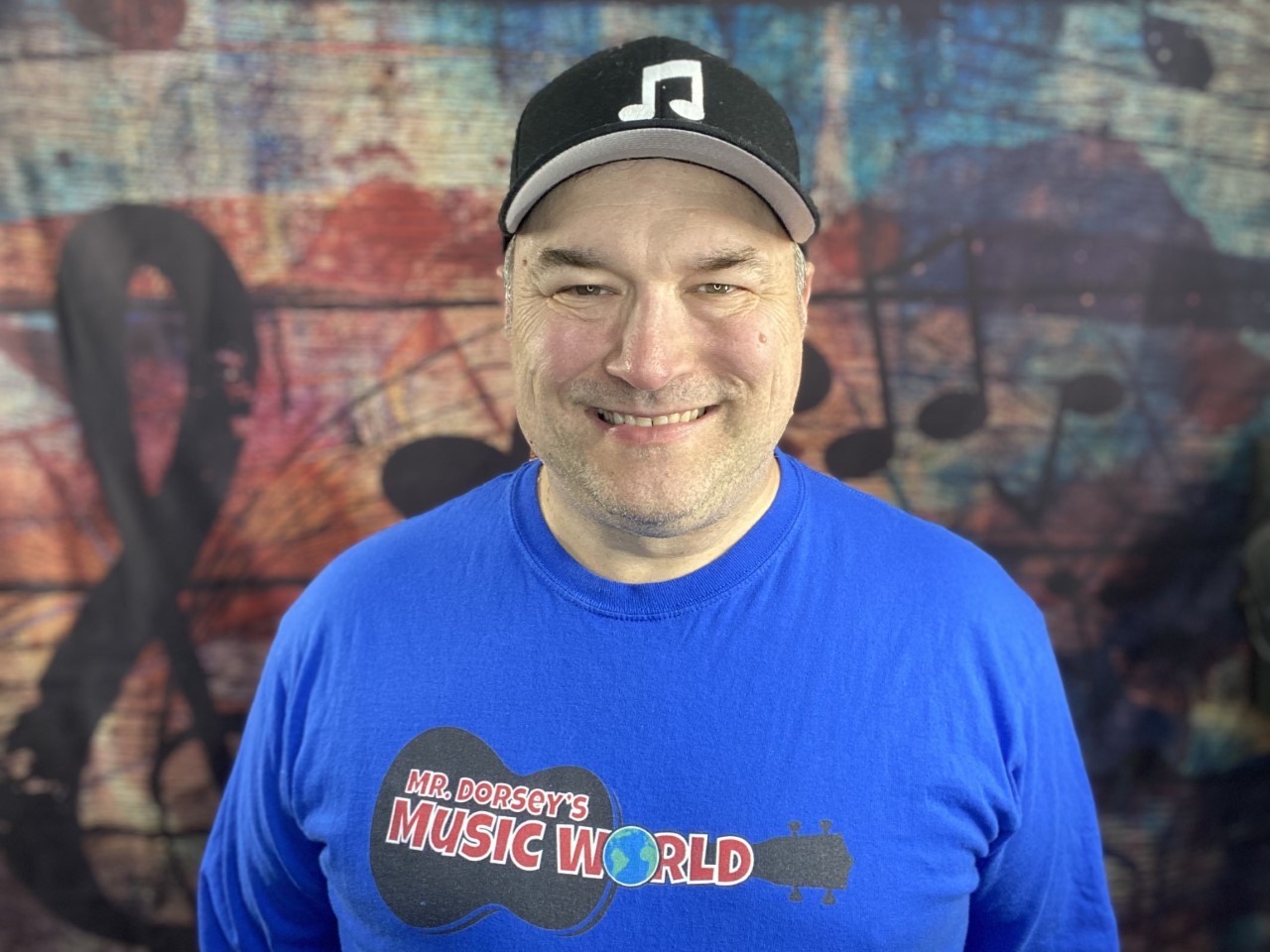 This is a photo of Seymour music teacher Kevin Dorsey, wearing a blue tshirt and black hat with a musical note on it, smiling at the camera.