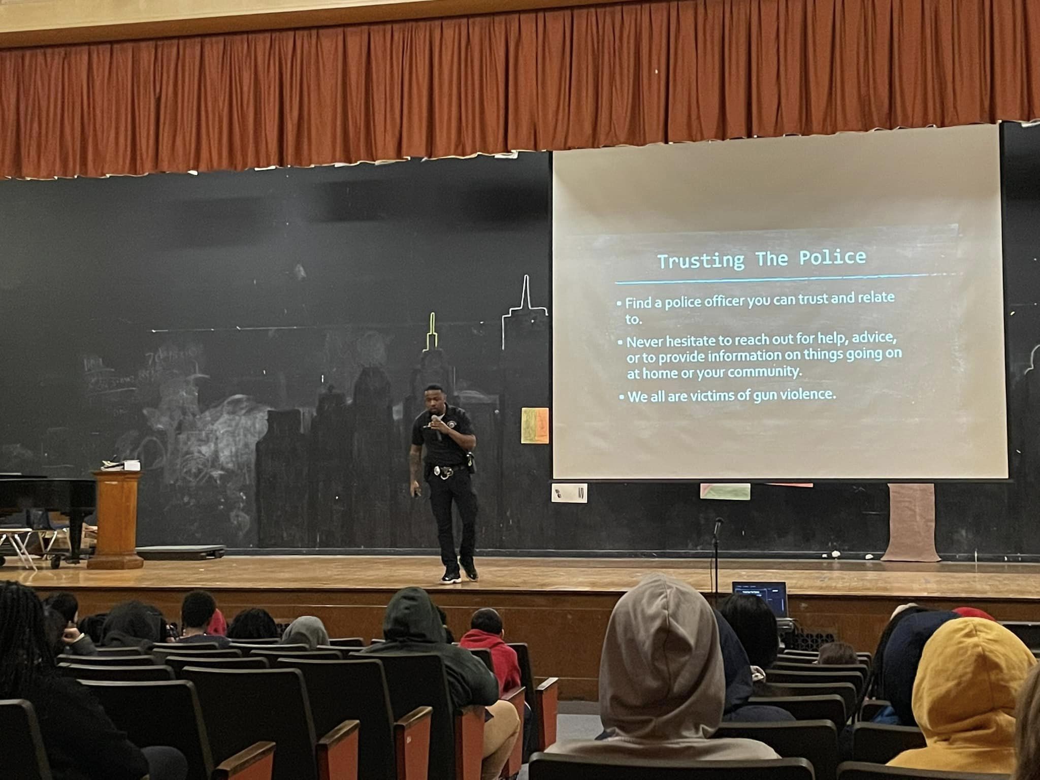 This is a photo of a police officer standing on a stage in a school auditorium speaking to students in the audience.
