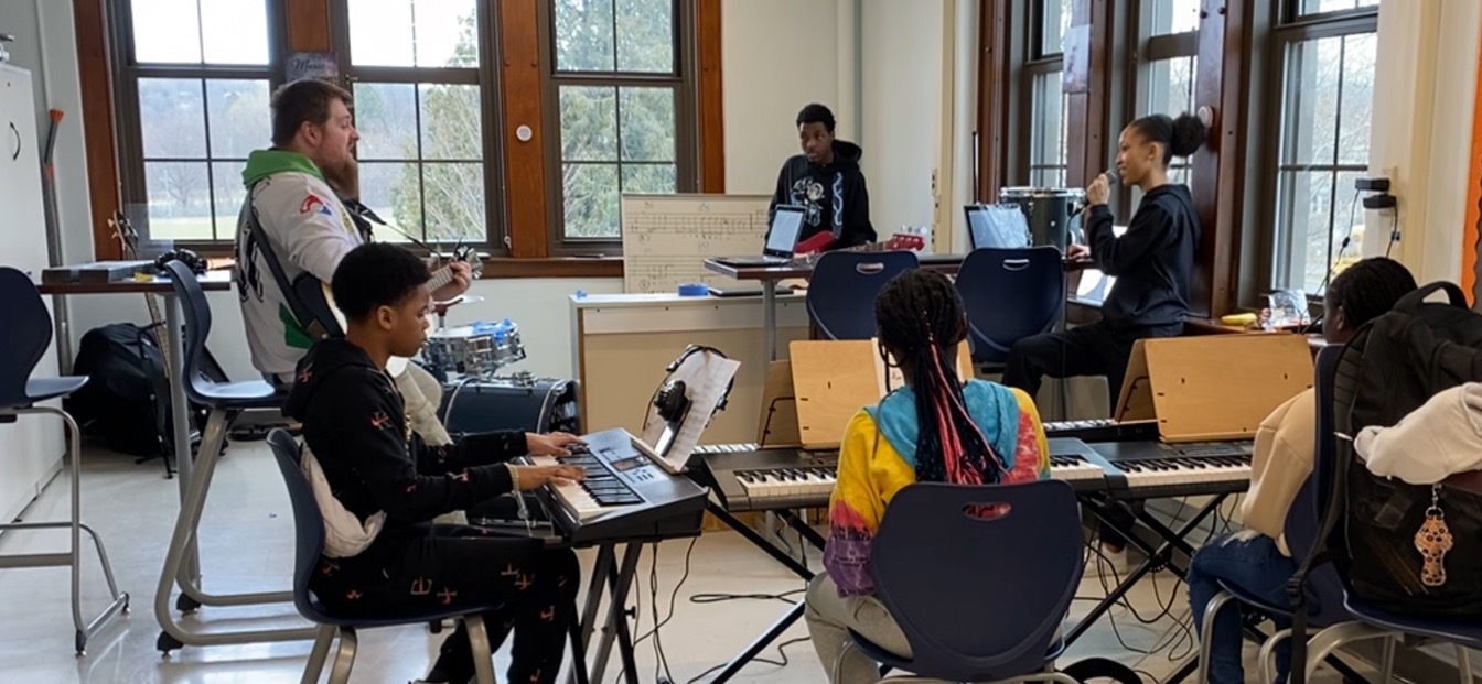 This is a photo of students in a music classroom, sitting in a circle each playing a musical instrument. 