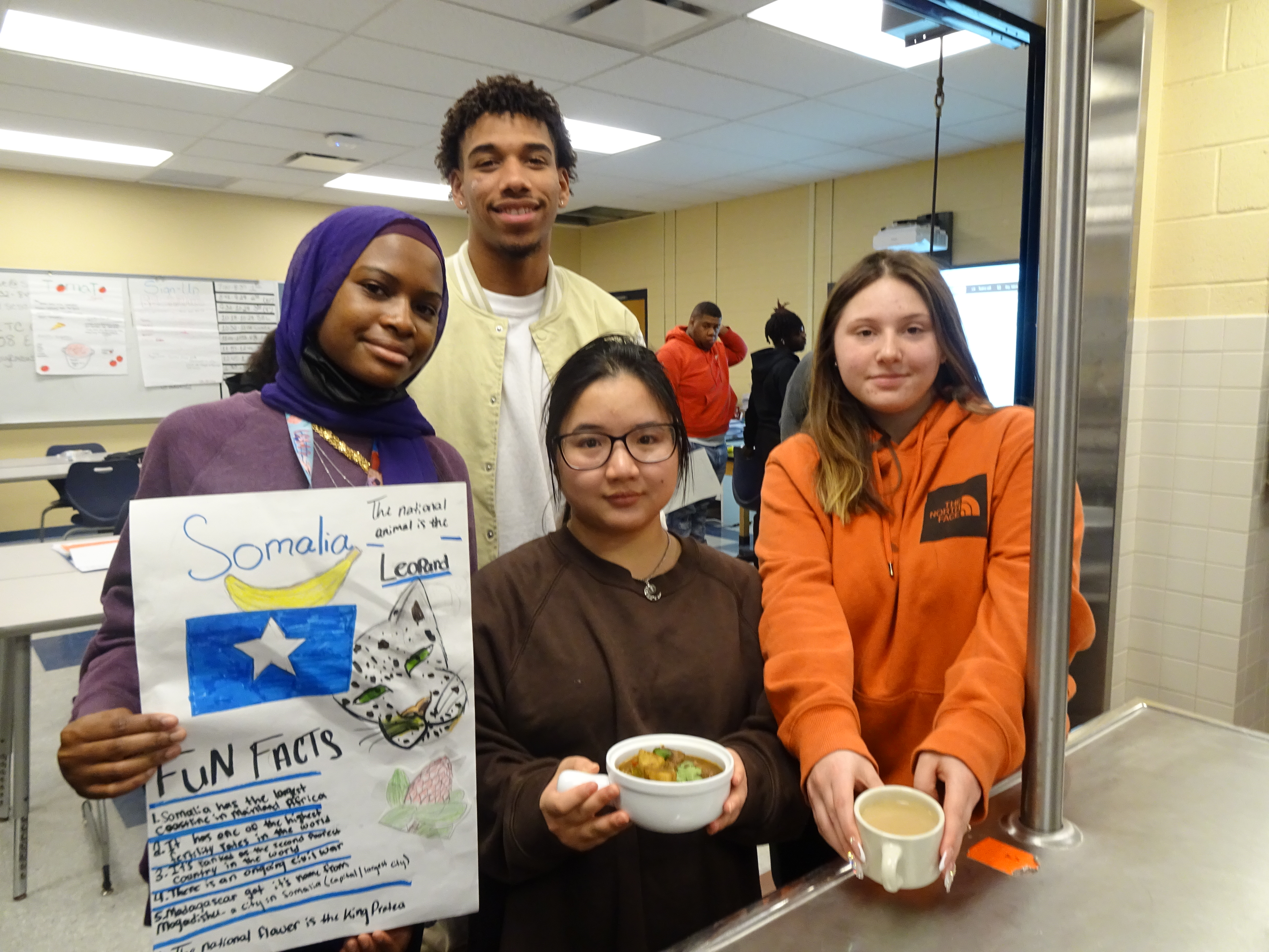 This is a photo of four ITC students standing in the kitchen in their school, holding a poster about Somalia and two dishes that they created from that nation.