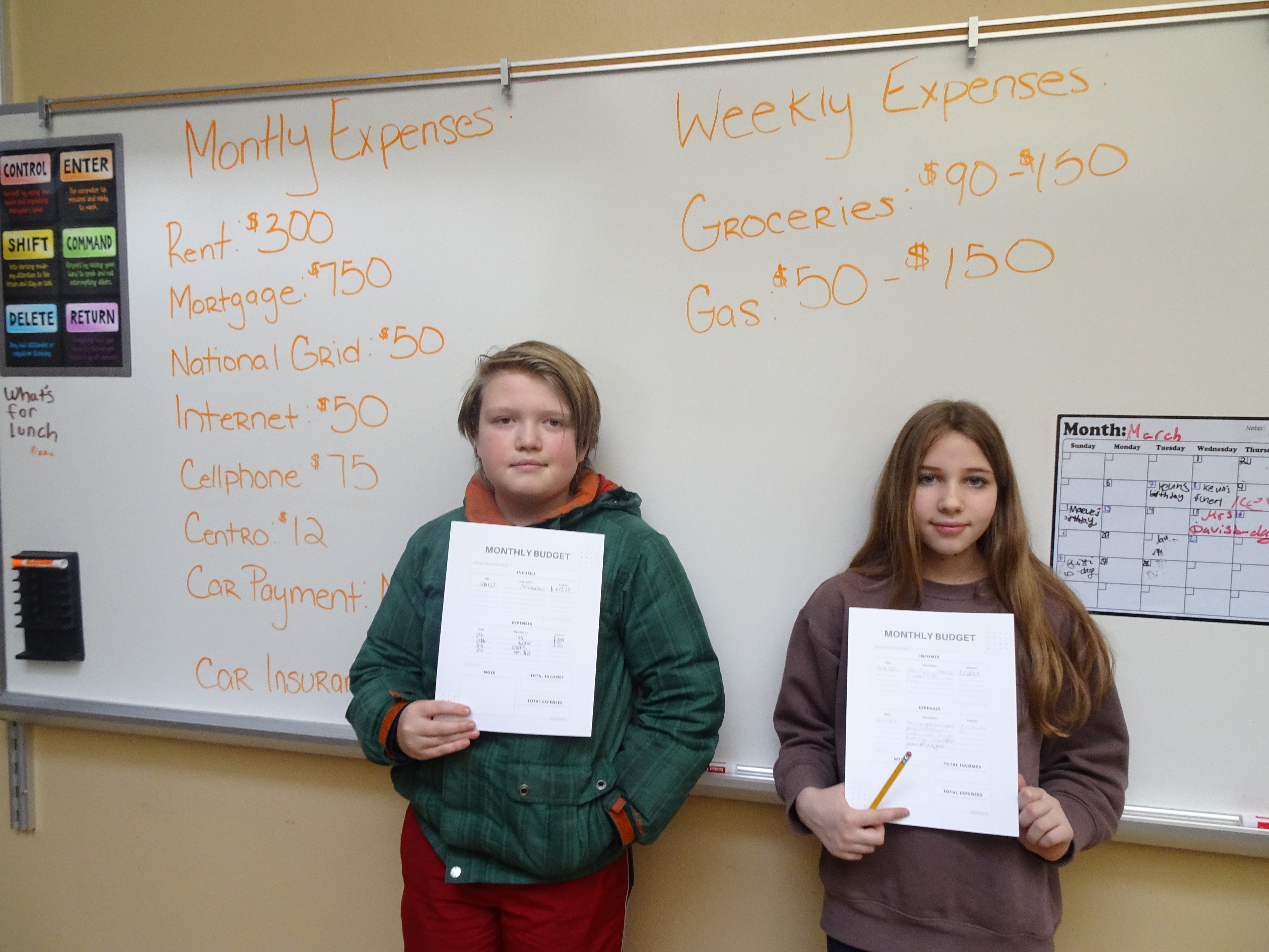 This is a photo of two Syracuse Latin students, each holding a sheet with their monthly budget, standing in front of a white board filled with a list of various household costs.