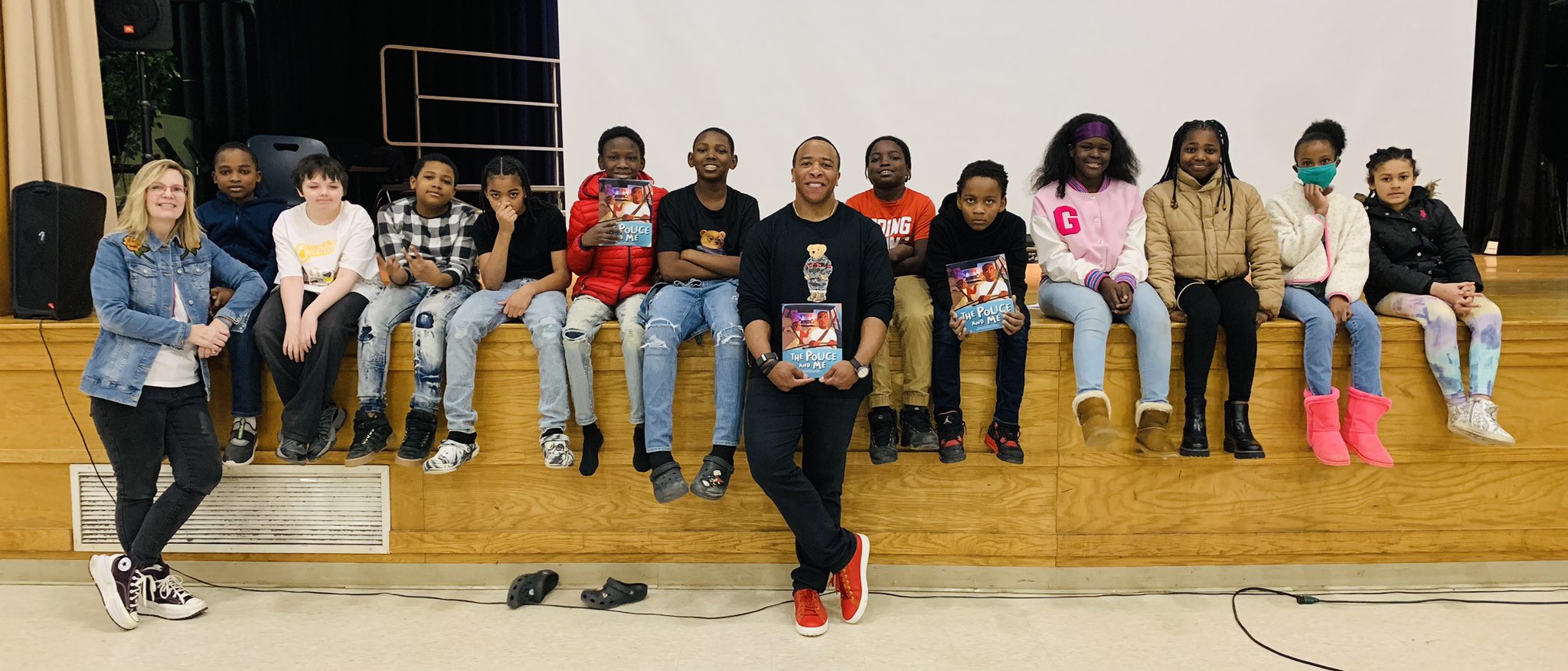 This is a photo of a group of 5th grade students, sitting on the edge of their school stage, with author Derrick Dotson in the center, smiling at the camera.