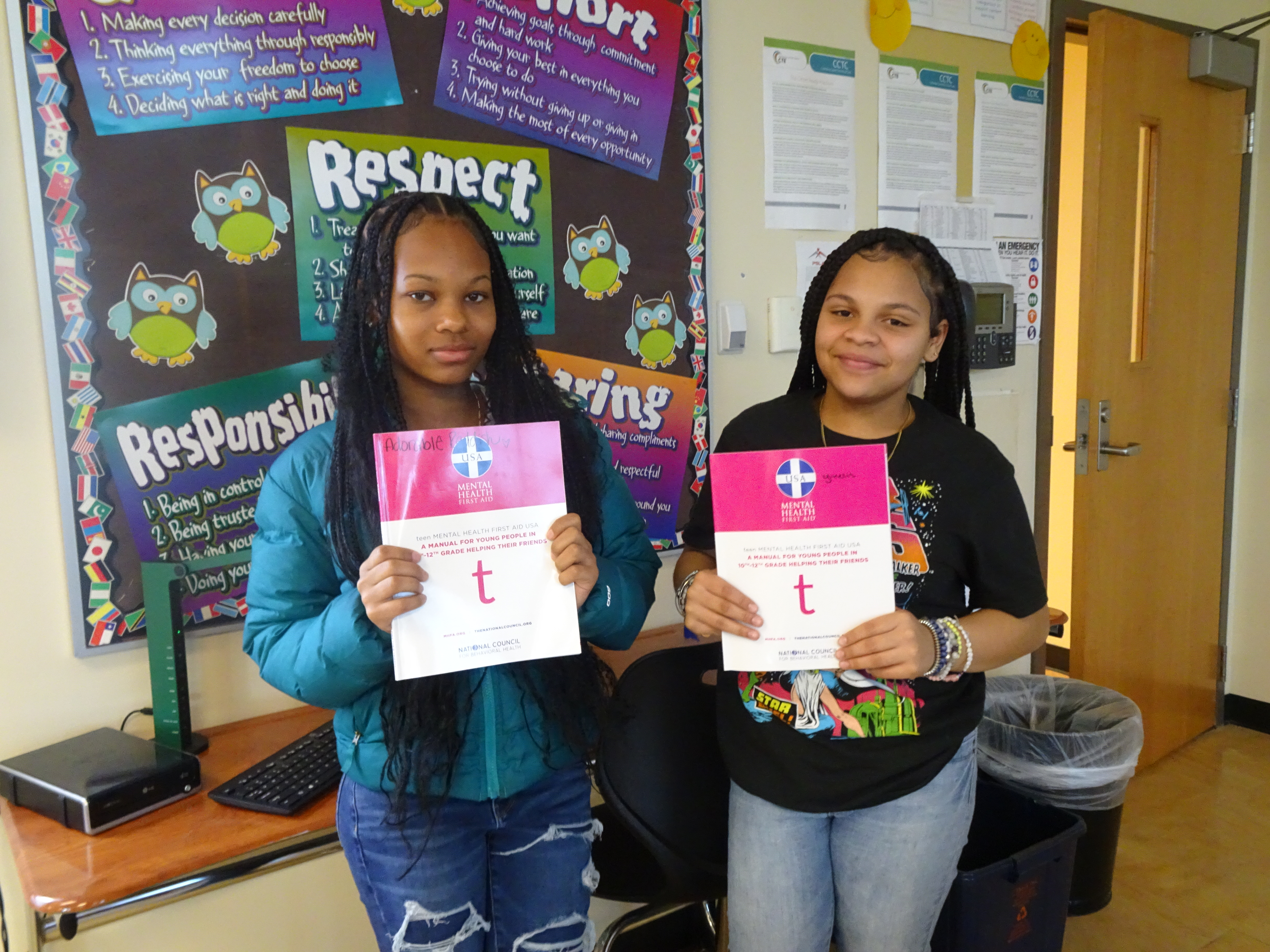 This is a photo of two female students standing in front of a bulletin board, each holding a mental health training booklet and smiling at the camera.