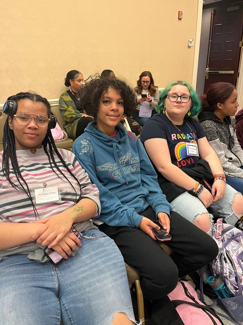 This is a photo of three Corcoran students, sitting in chairs in a ballroom and smiling for the camera as they await the start of a motivational speaker's session.