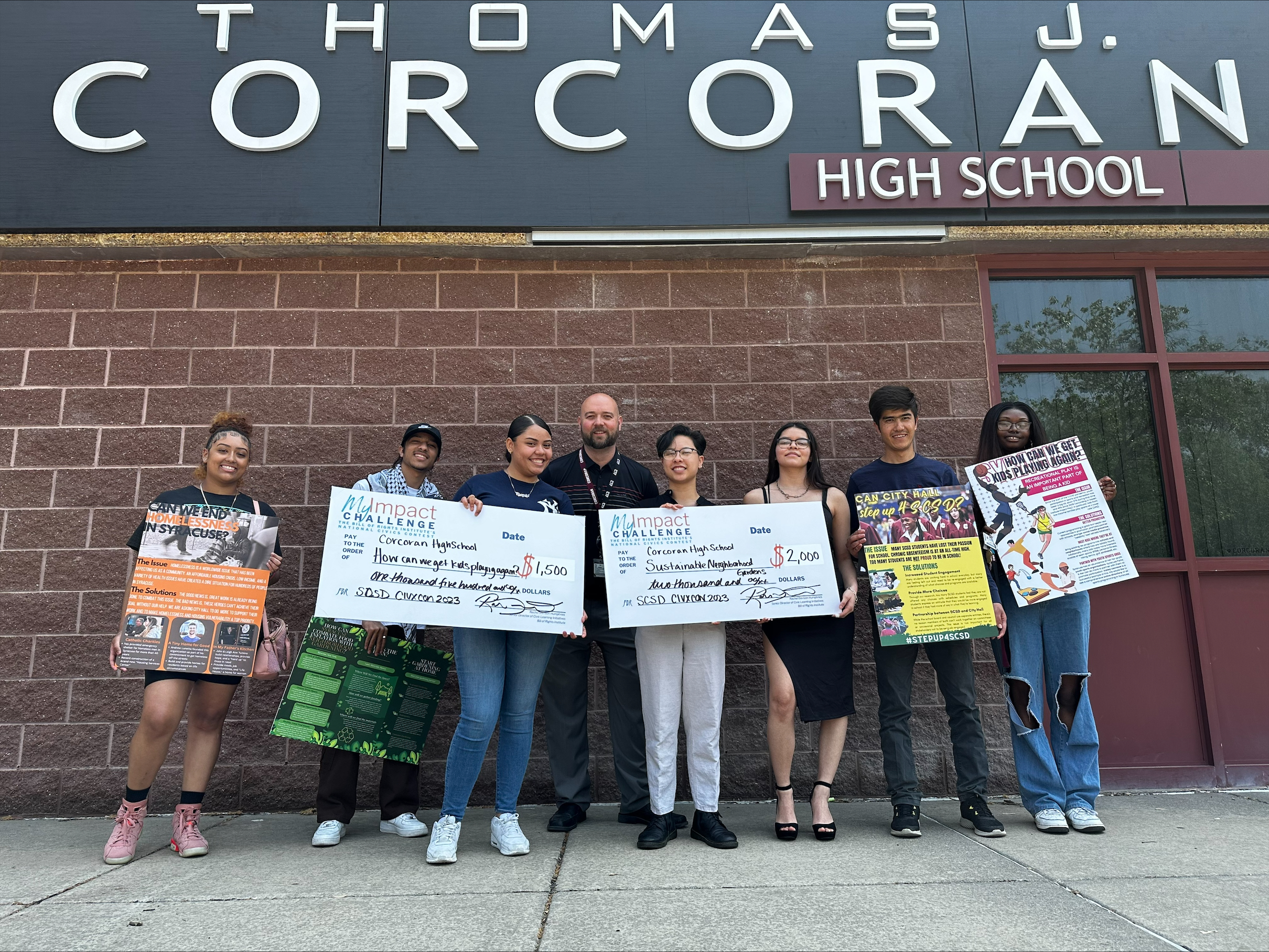 This is a photo of a group of Corcoran high school students standing outside their school building holding posters and checks they received through the Syracuse Youth Advisory Council.
