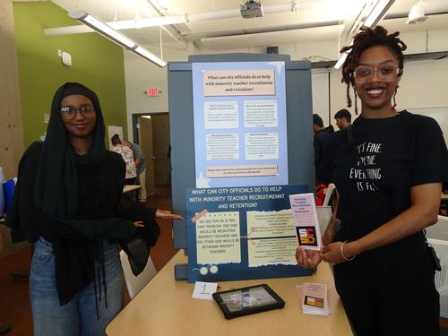 This is a photo of two students standing on either side of a poster they created, smiling at the camera. 