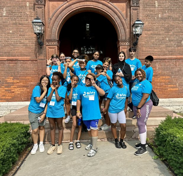 This is a photo of a group of GEAR UP students, all wearing matching blue tshirts and smiling outside a building on the Wells College campus.