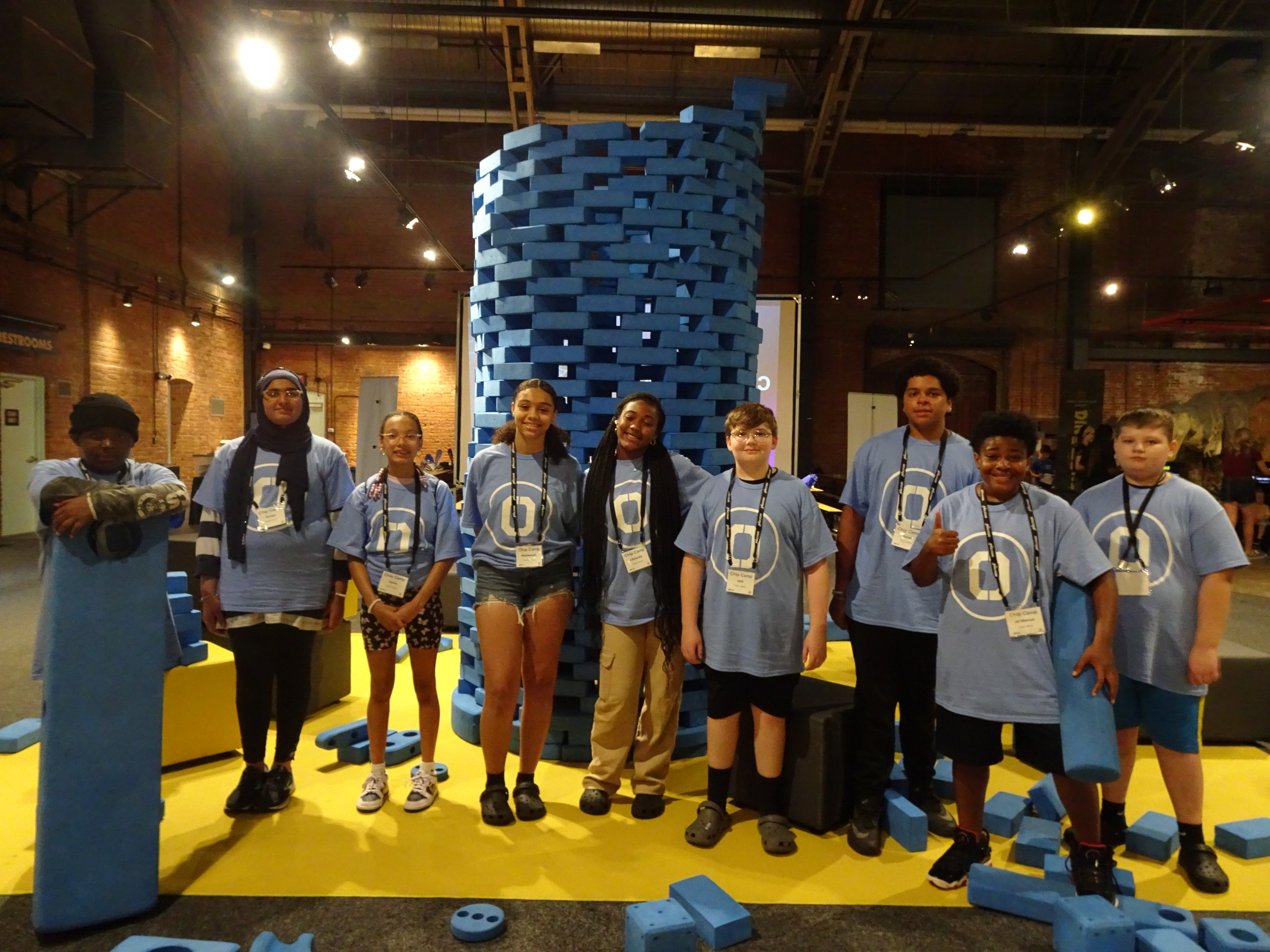 This is a photo of a group of students standing in front of a tall tower of blocks at the MOST, smiling at the camera.