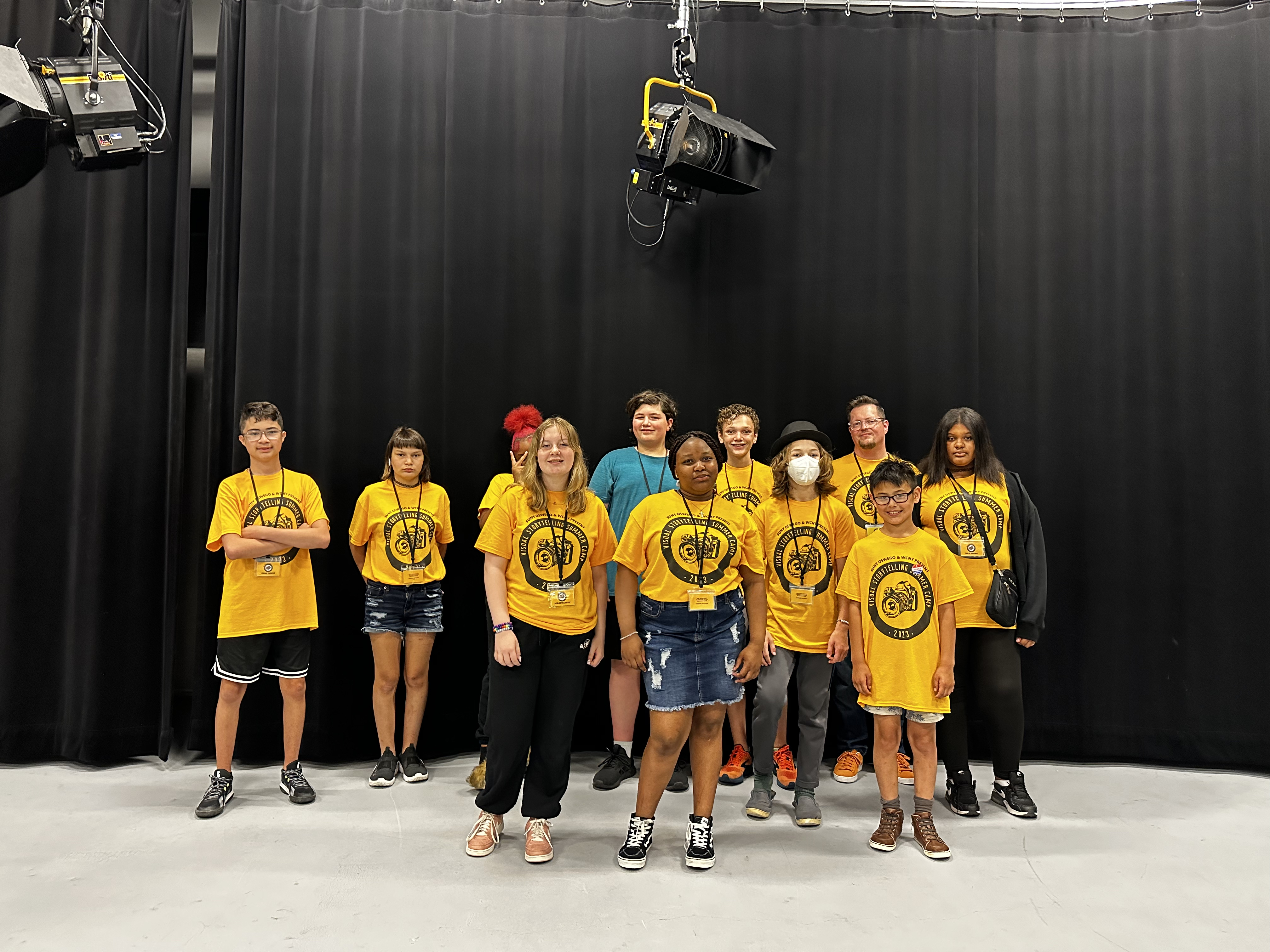 This is a photo of a group of students standing in a WCNY TV studio wearing matching yellow tshirts and smiling at the camera.