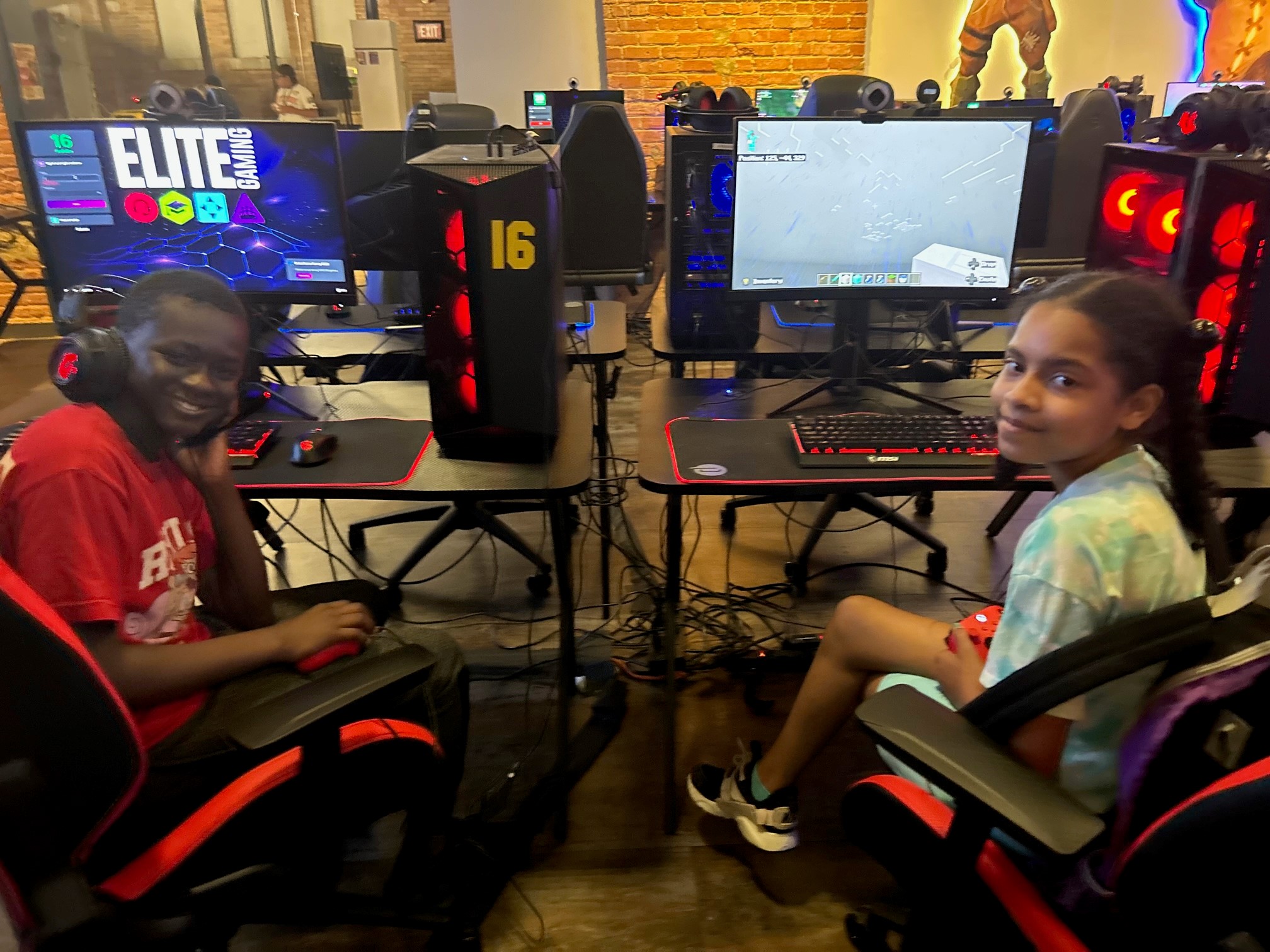 This is a photo of two students, each sitting at a computer gaming center and smiling at the camera.