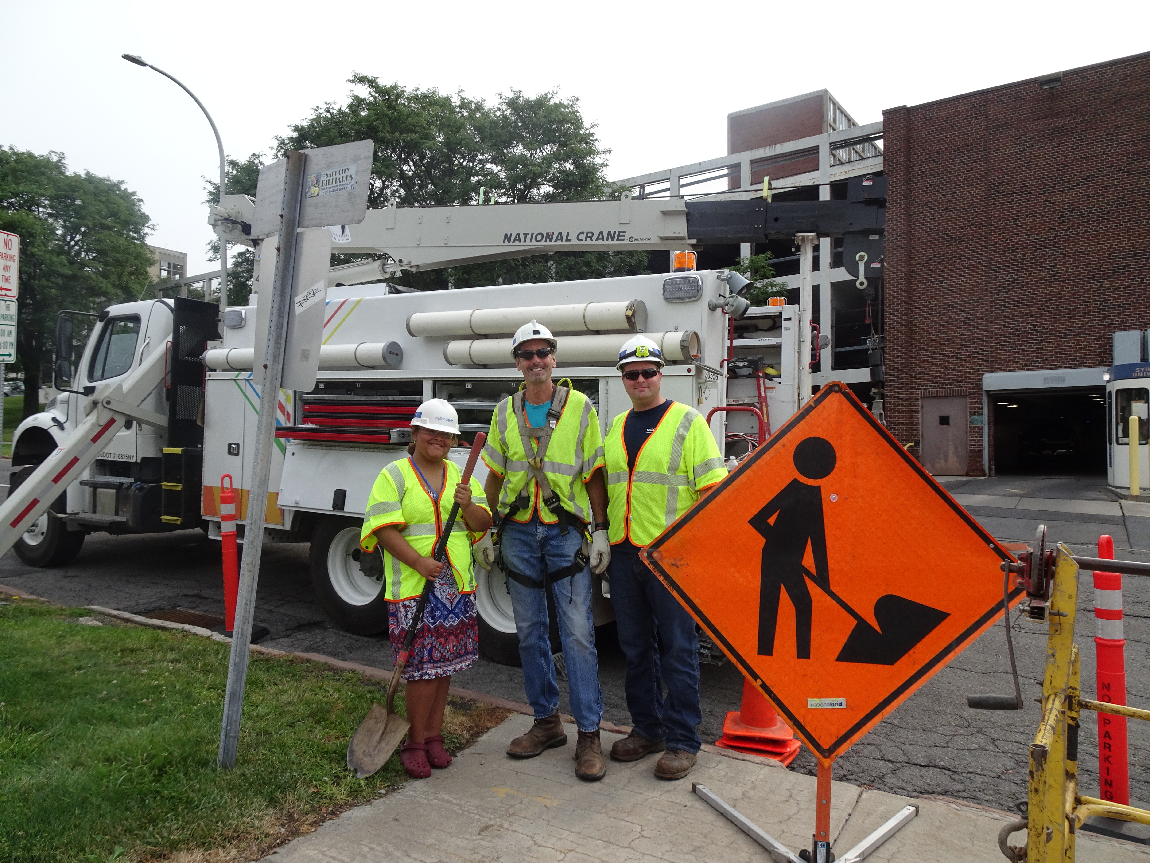 This is a photo of a student standing in a safety vest and hard hat alongside two National Grid employees, in front of a National Grid truck.