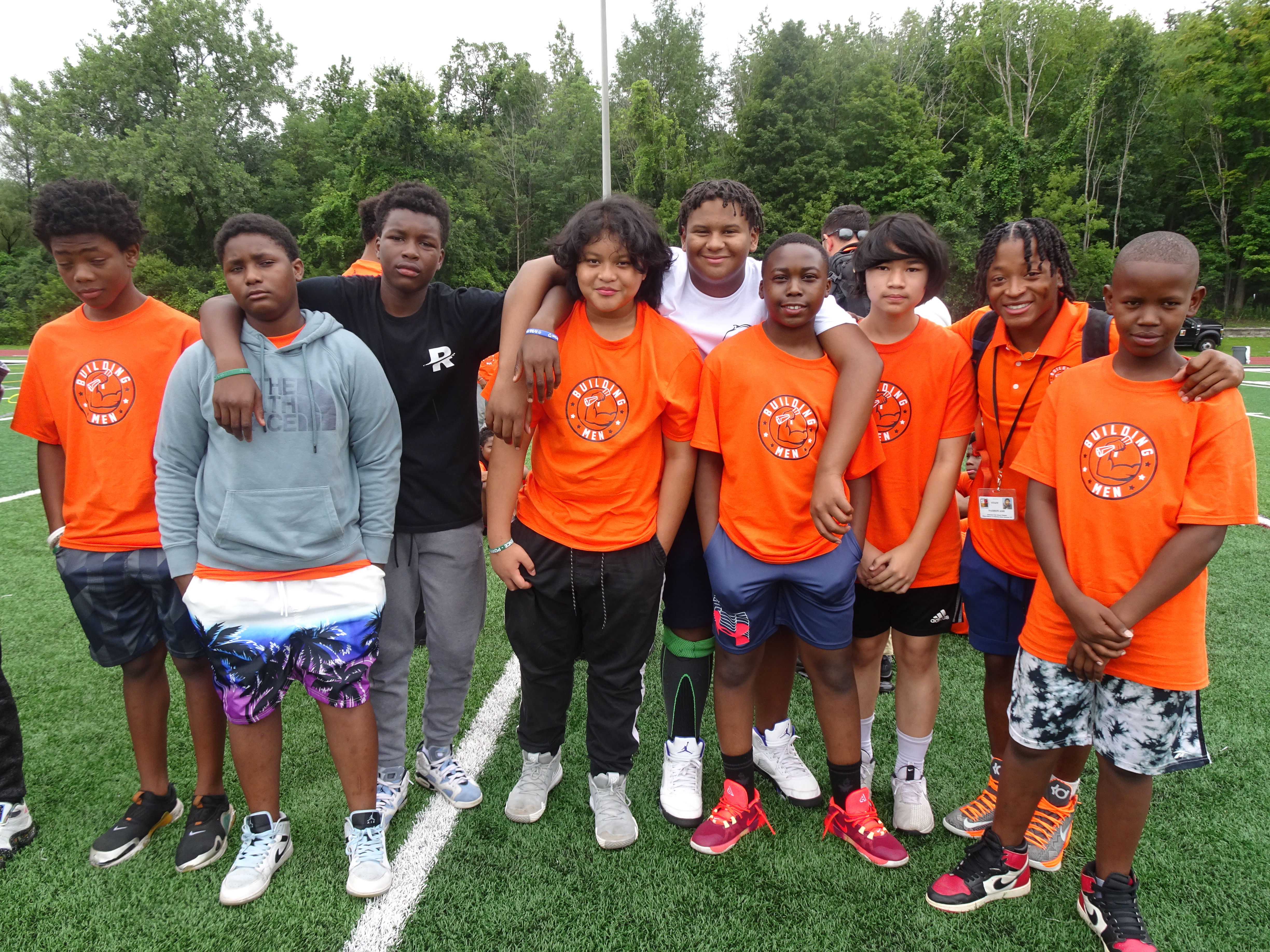 This is a photo of a group of Building Men Progra, middle school students, wearing matching orange shirts, standing in a huddle and smiling at the camera.