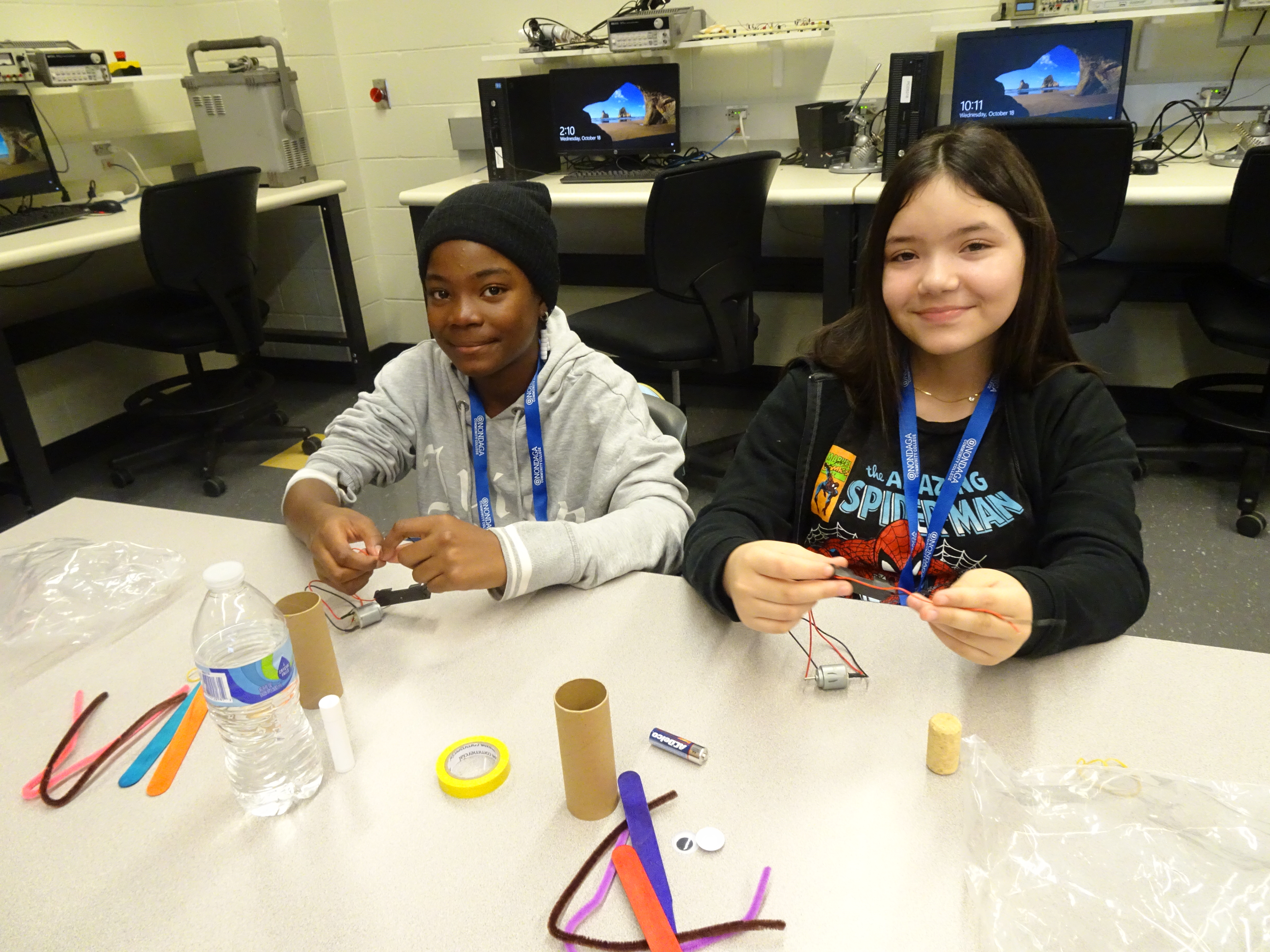 This is a photo of two middle school girls sitting at a desk working on building wiggle robots and smiling at the camera.
